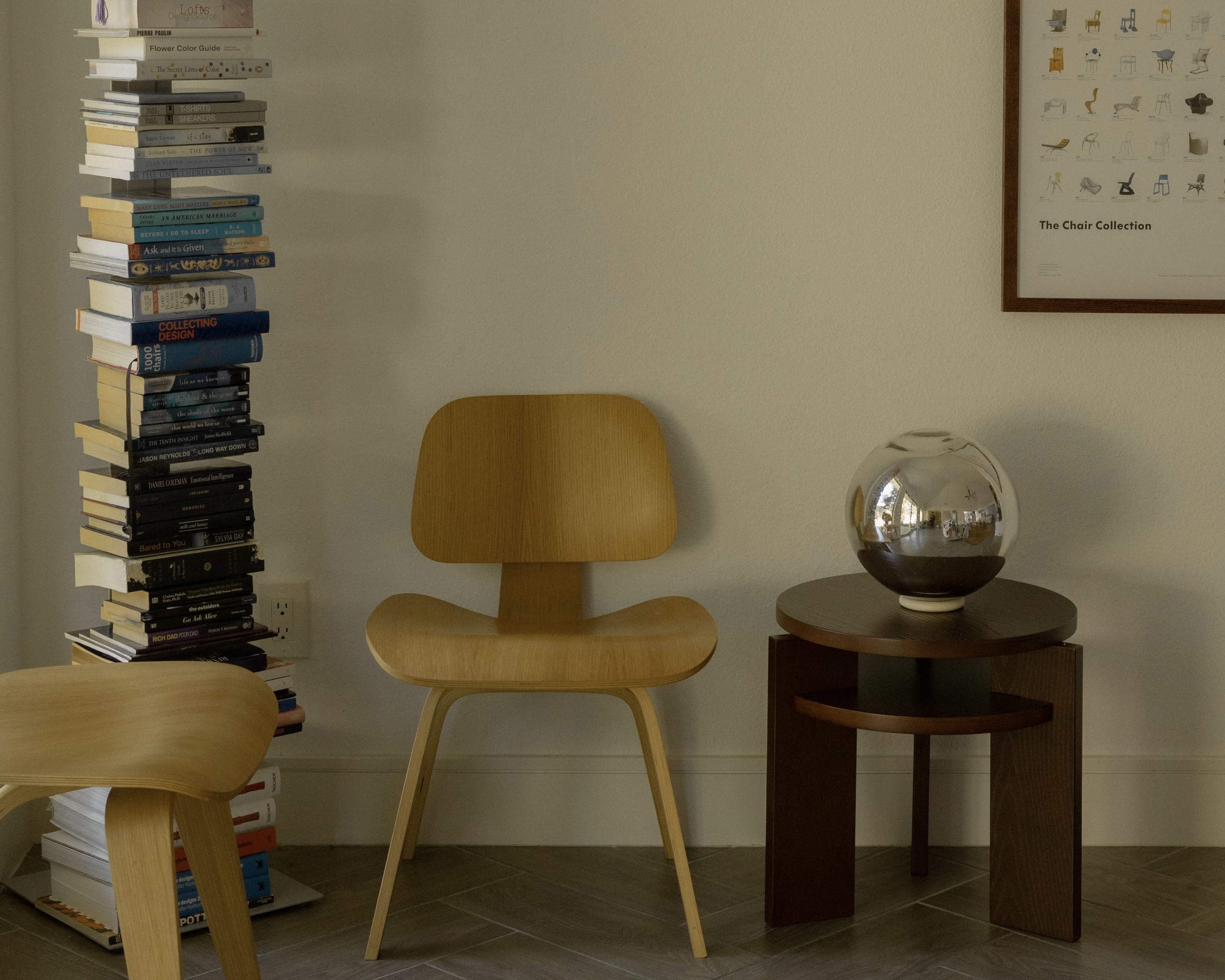 A wooden chair is positioned beside a stacked column of books and a round table with a decorative glass object on top.