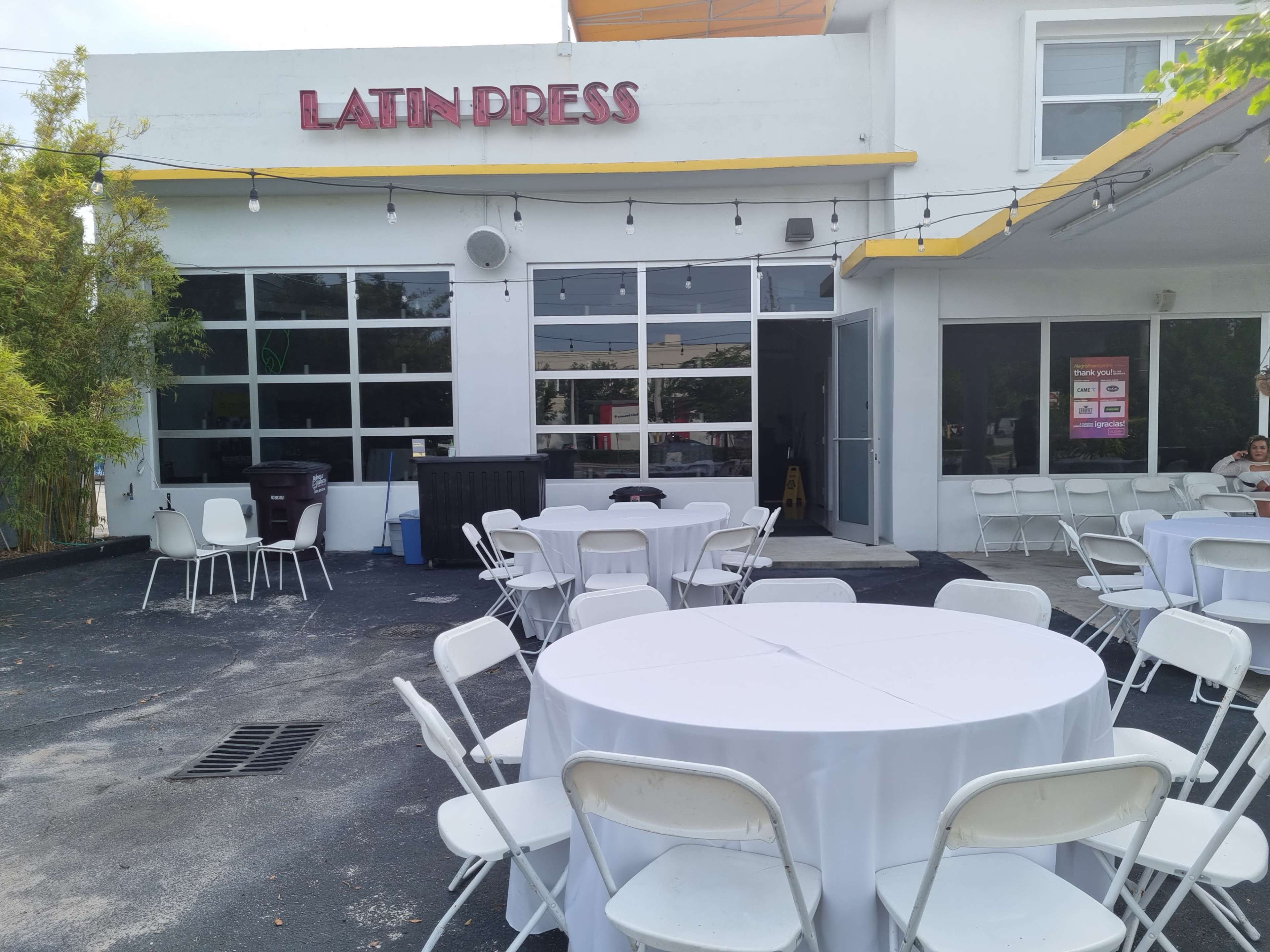 The exterior of a building labeled "LATIN PRESS," featuring empty round tables and white chairs arranged outside on a pavement.