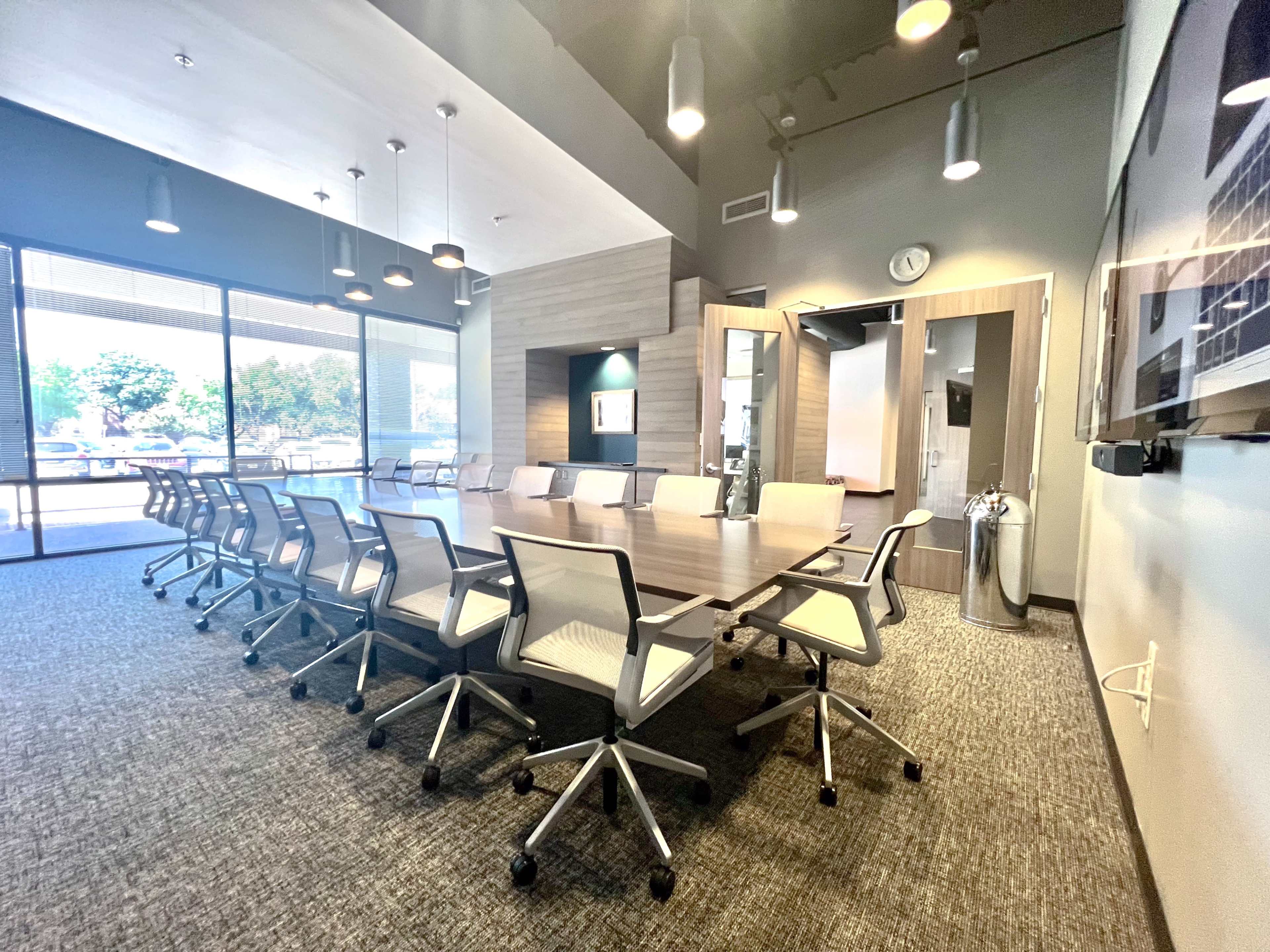 A large conference room features a long wooden table surrounded by ergonomic chairs, with large windows allowing natural light to fill the space.