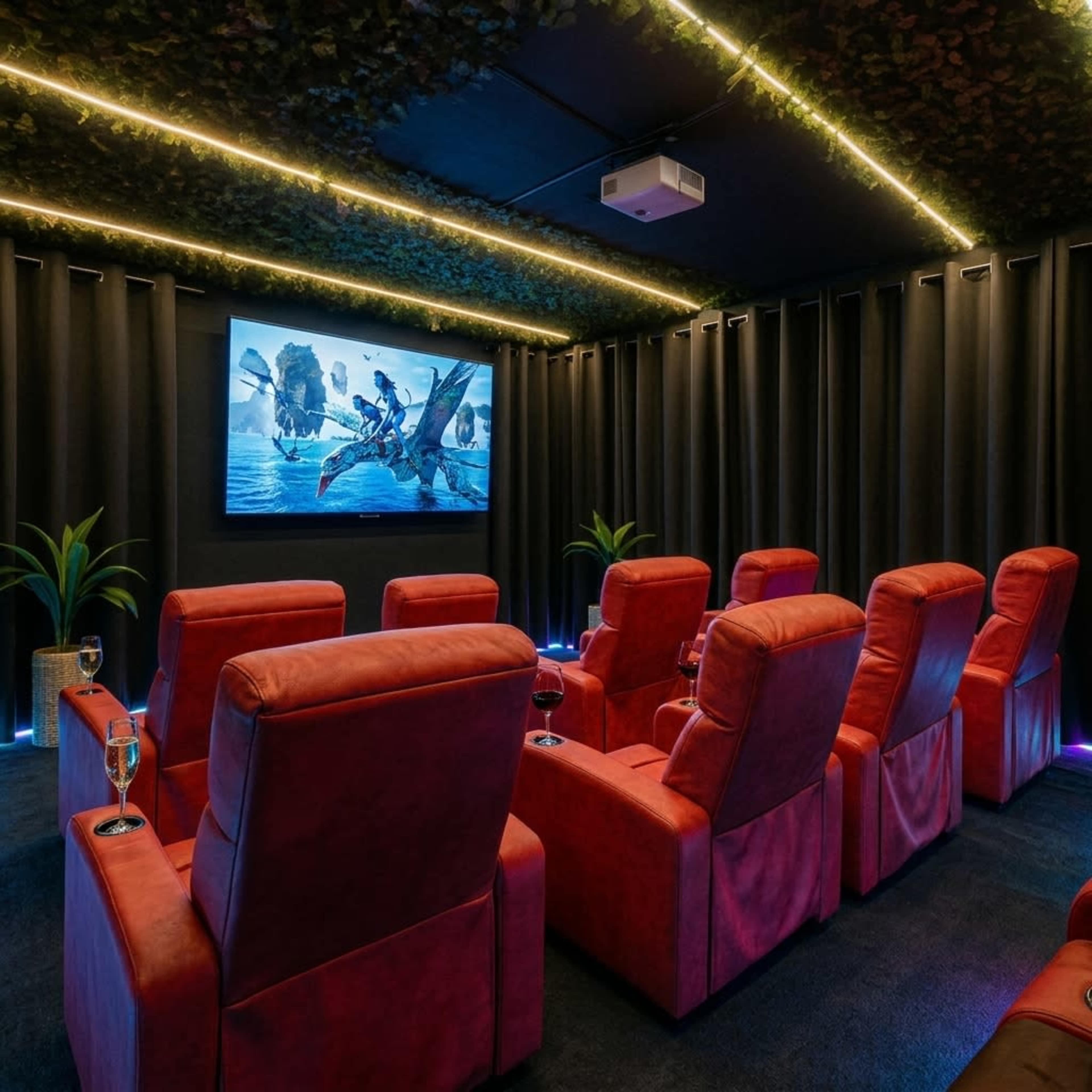 A home theater features red reclining chairs arranged in rows facing a wall-mounted screen, with overhead lights and potted plants adding decor.