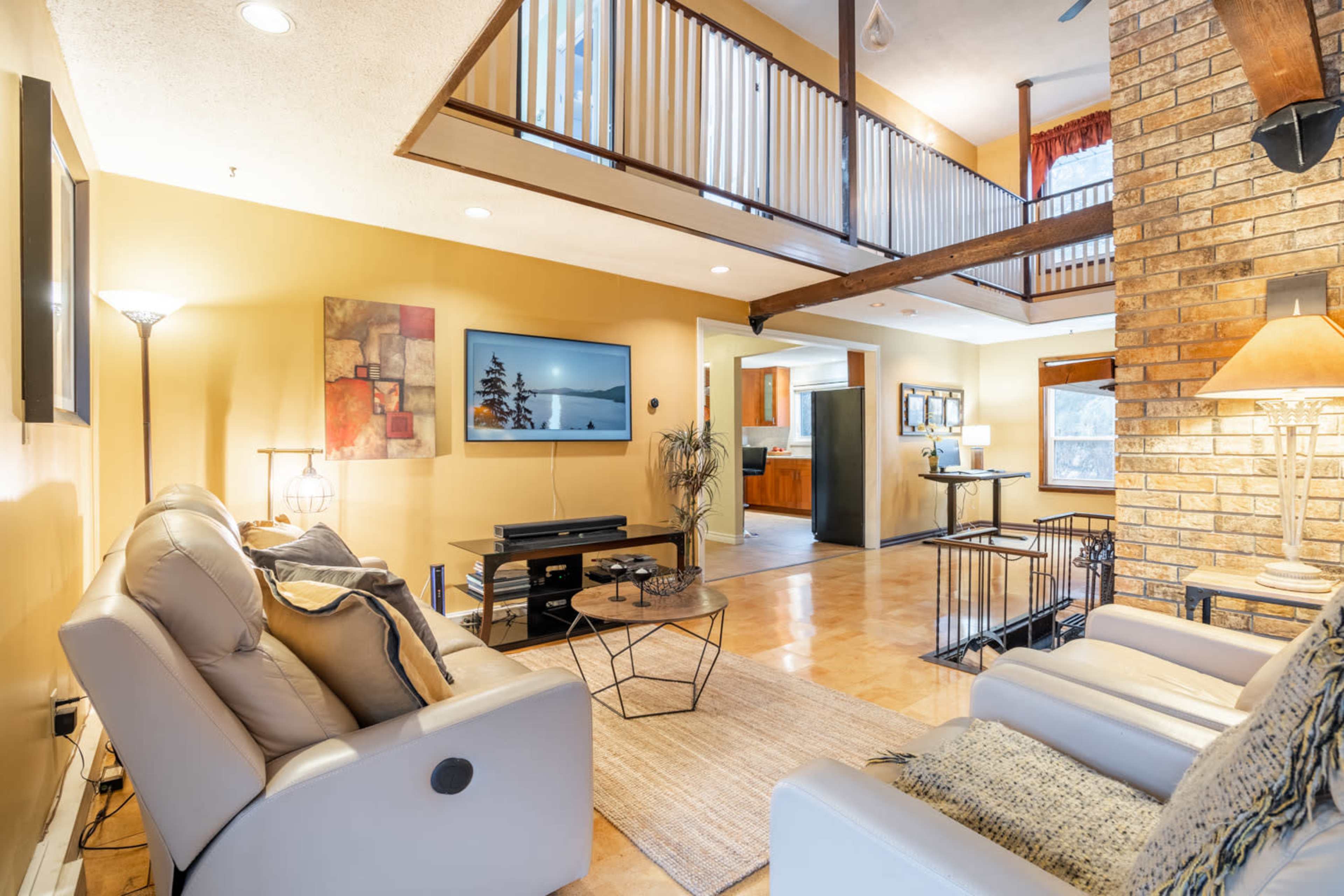 The image shows a spacious living area with a seating arrangement, a television mounted on the wall, and a view of the upper balcony and kitchen area.