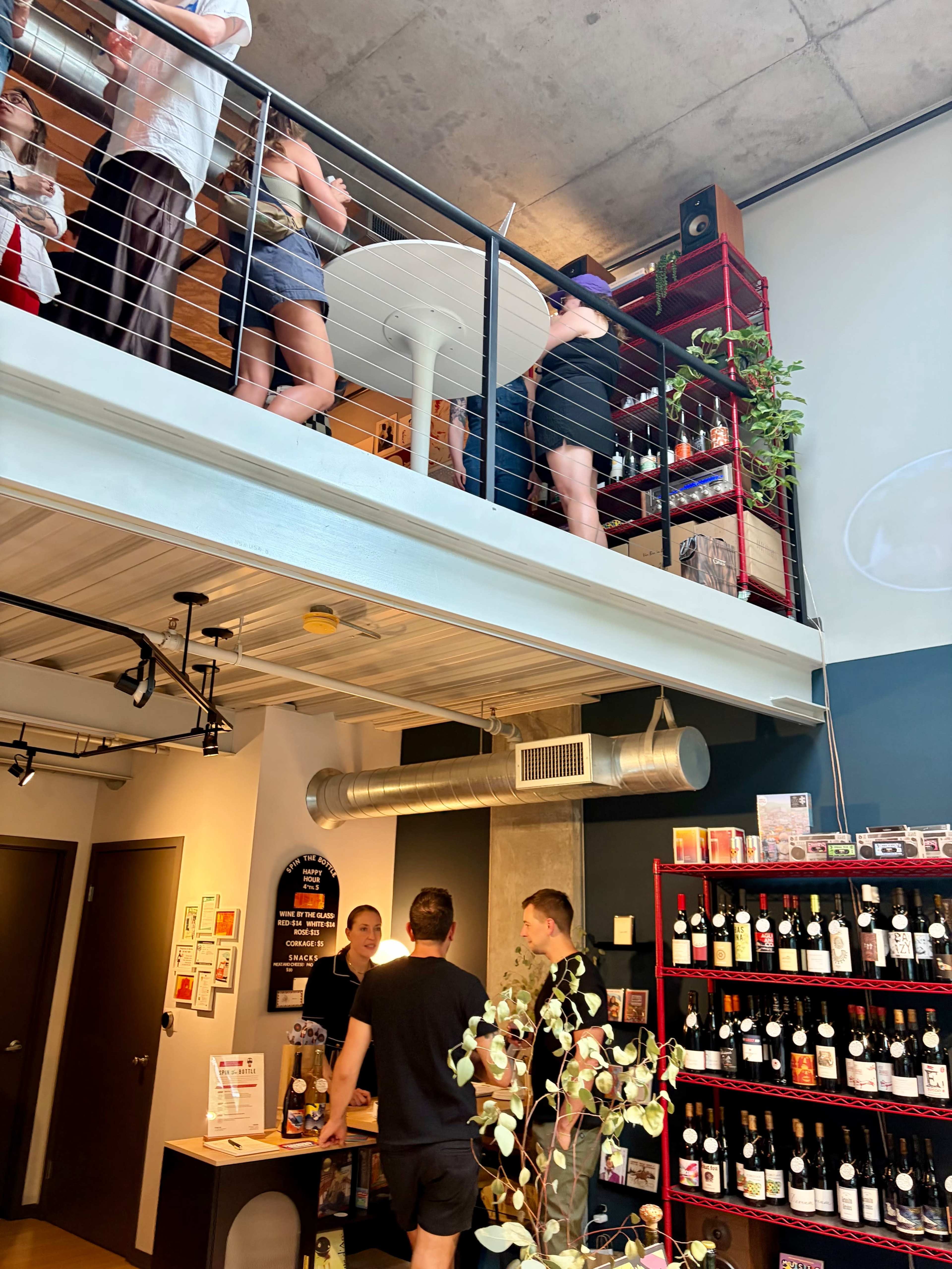 A group of people stands on a balcony overlooking a bar area filled with shelves of wine bottles and a few patrons at the counter.