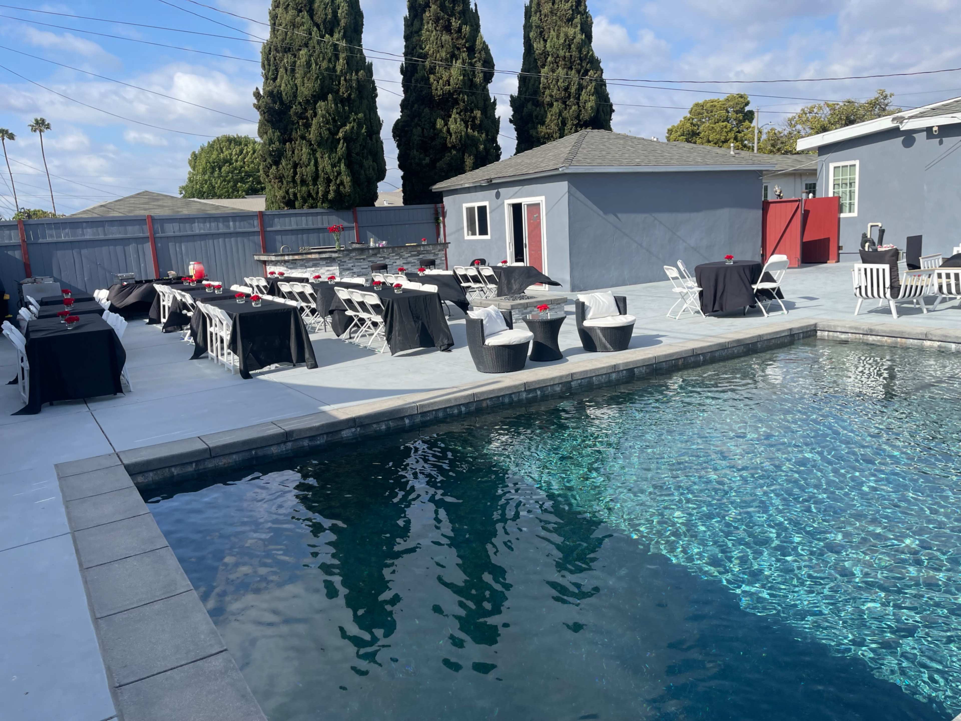 The image shows a pool area with several tables and chairs arranged around it, a house in the background, and tall trees nearby.