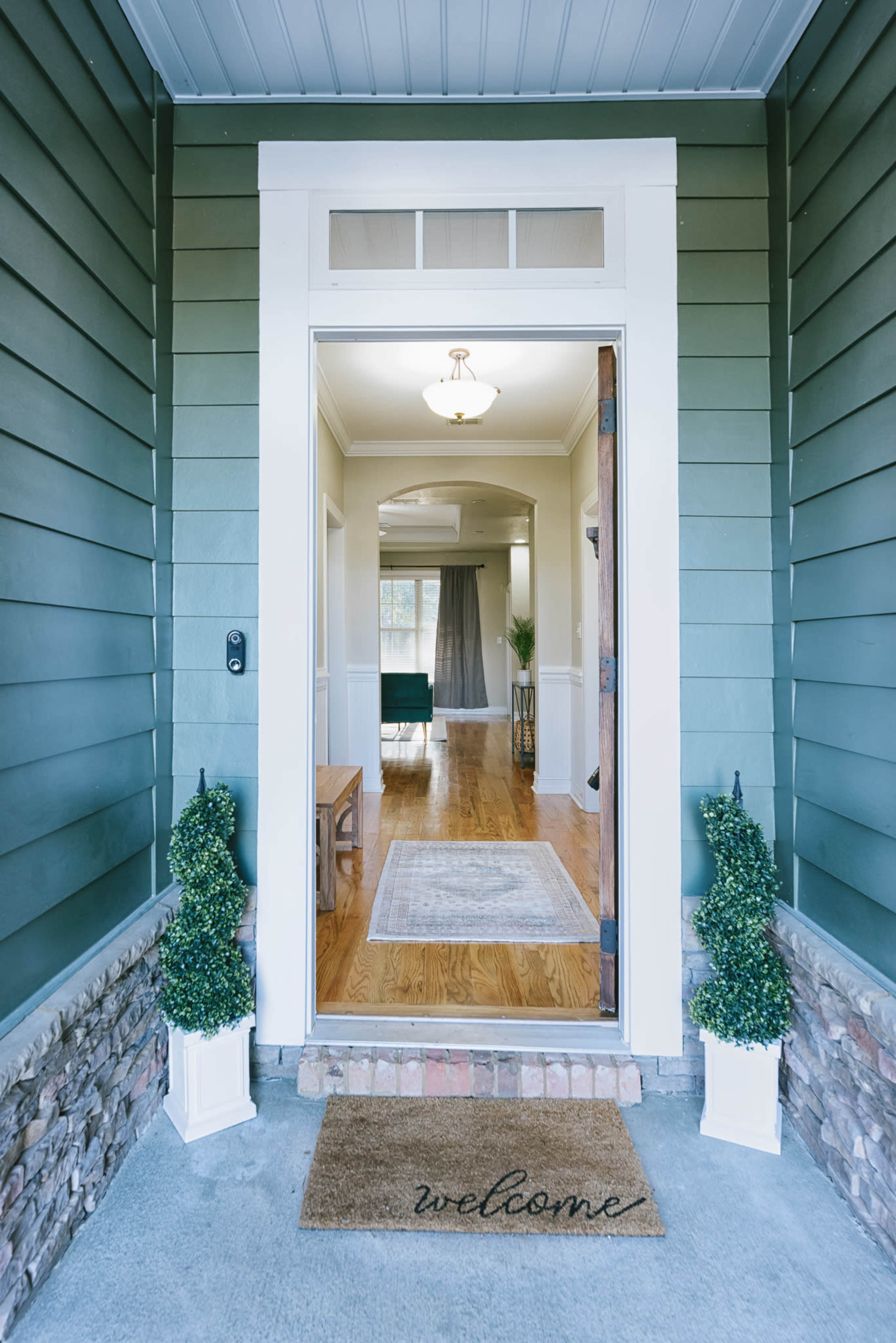 The entrance of a home with a welcome mat and potted plants on either side of the doorway, leading into a well-lit living area.