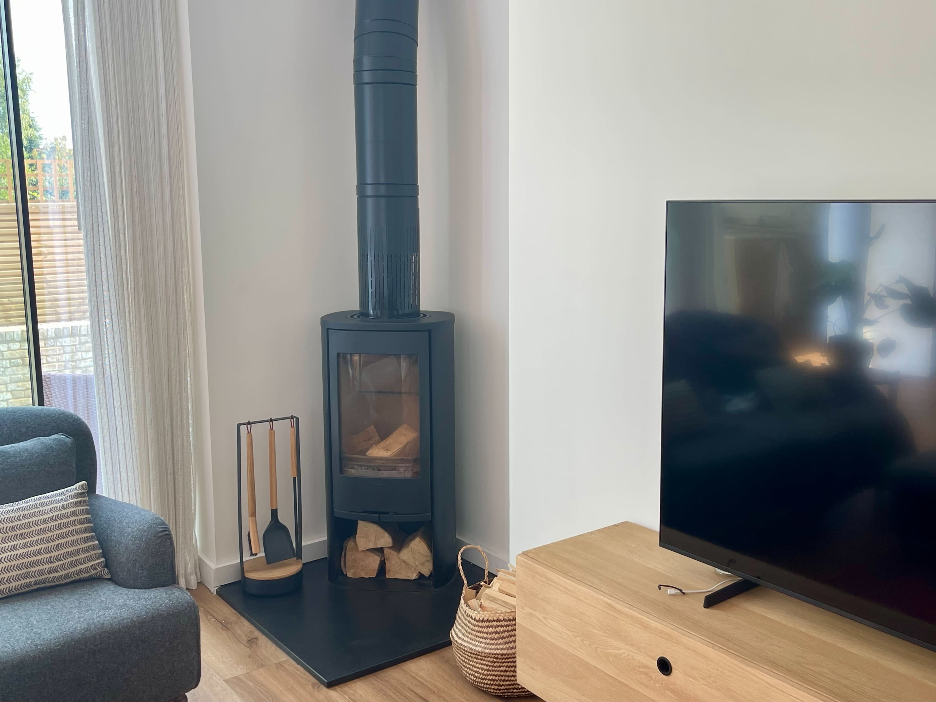 The image shows a modern living room with a black wood stove next to a TV and a couch, along with a basket of logs.