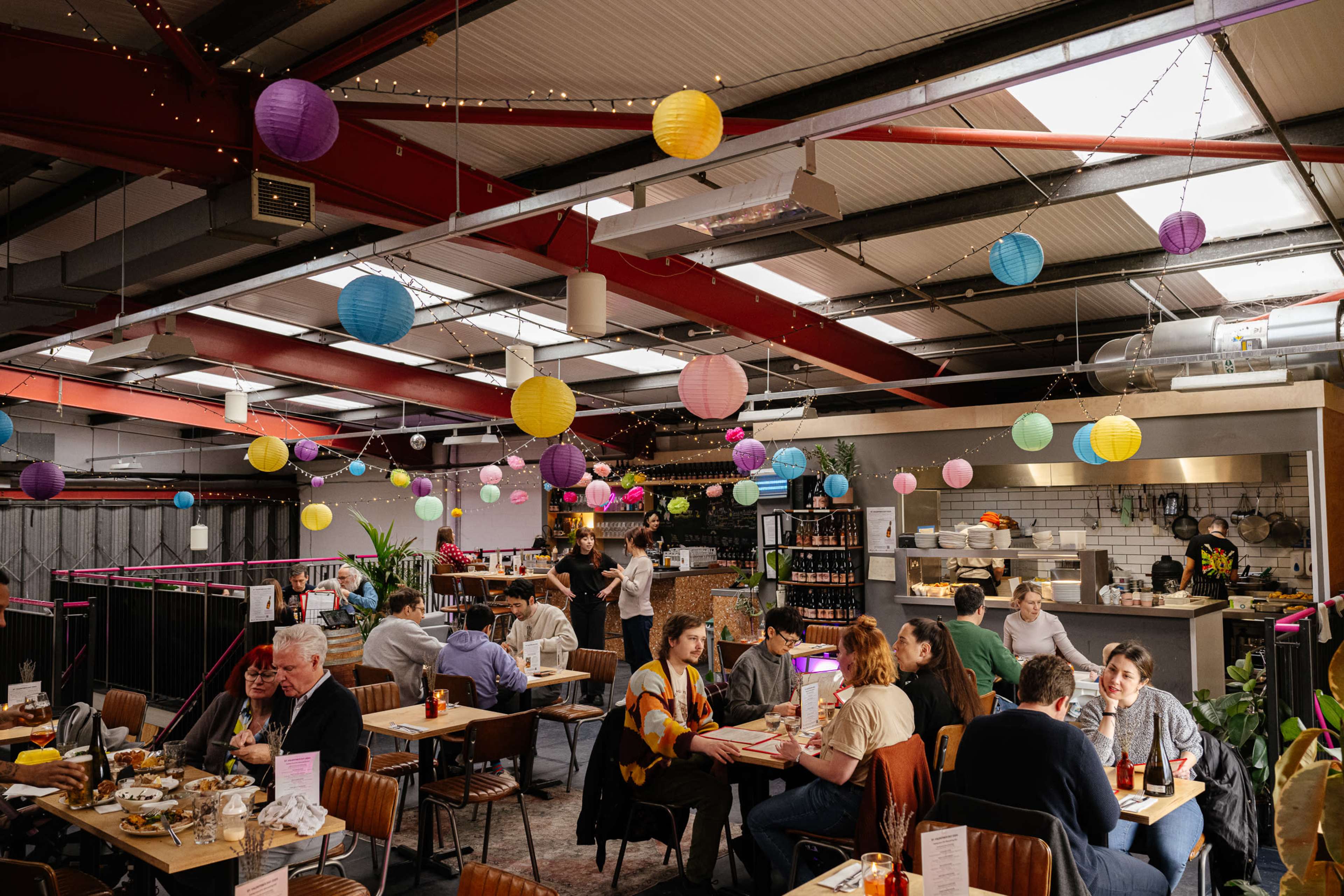 A lively restaurant features colorful paper lanterns hanging from the ceiling, with diners seated at tables enjoying their meals and staff serving food in an open kitchen area.