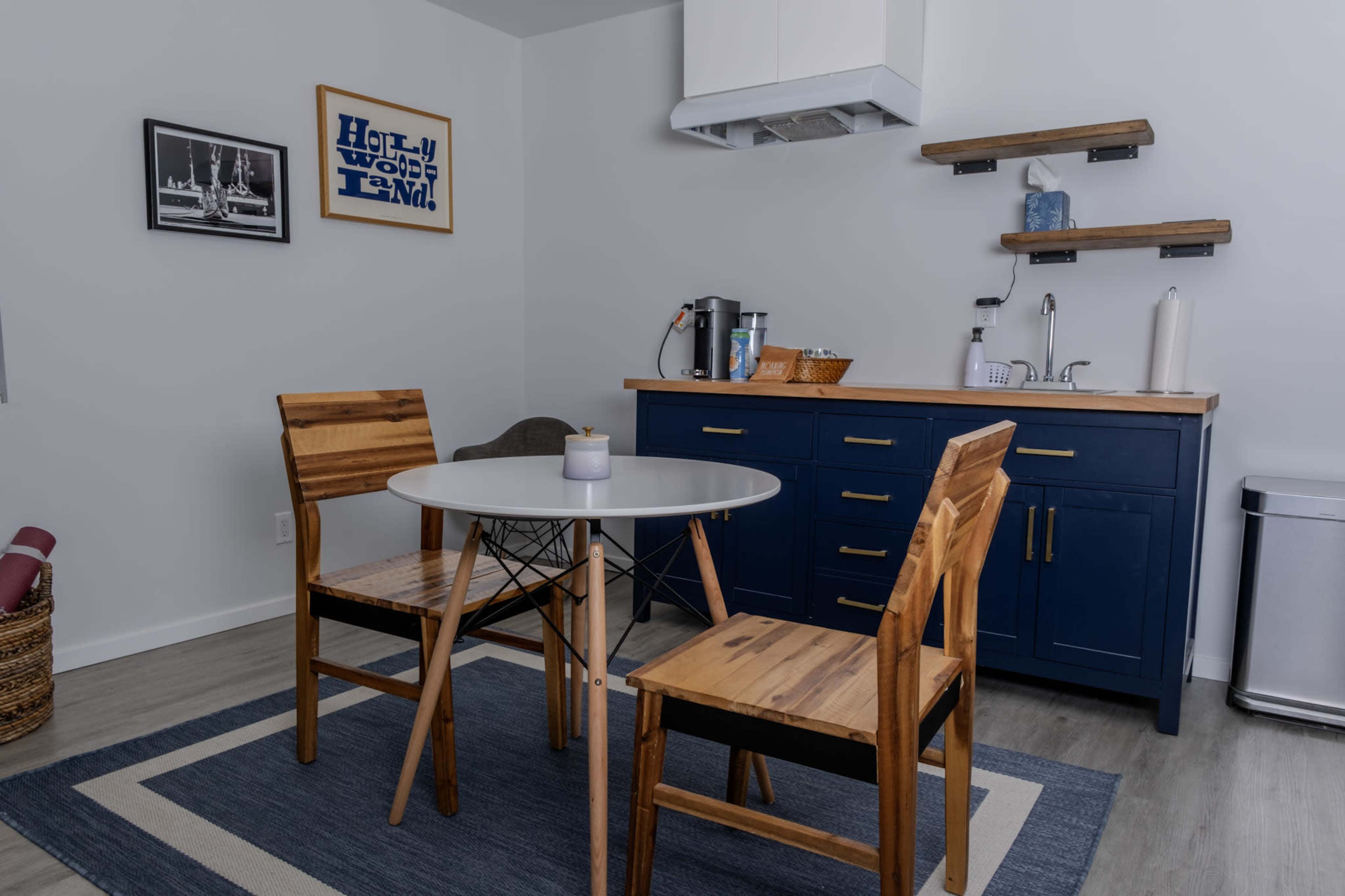 The image shows a modern kitchen area with a circular table and two wooden chairs, a dark blue cabinet, and a small appliance counter.