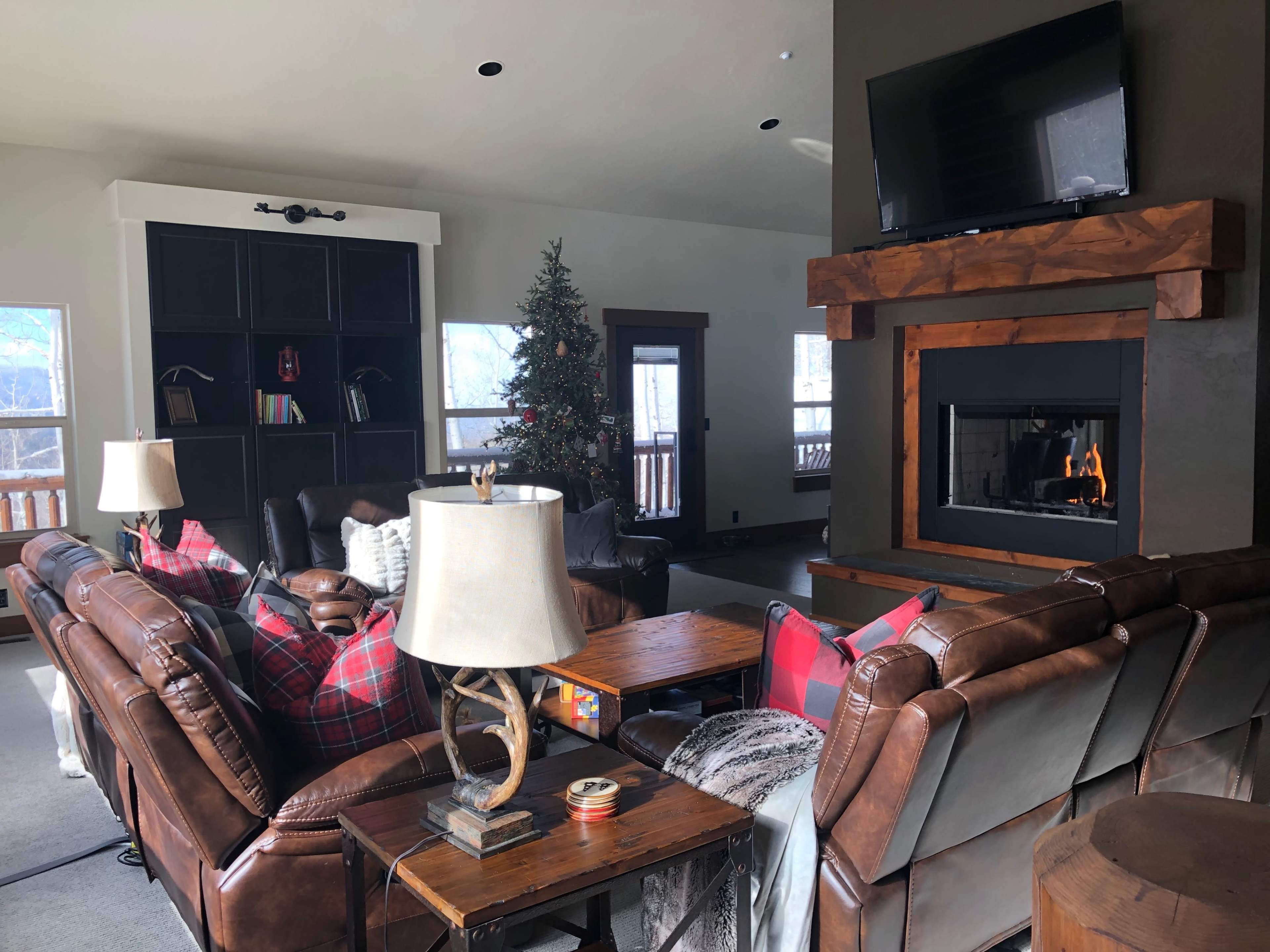 A spacious living room with brown leather sofas, a wooden coffee table, a decorated Christmas tree, and a fireplace with a flat-screen TV above it.