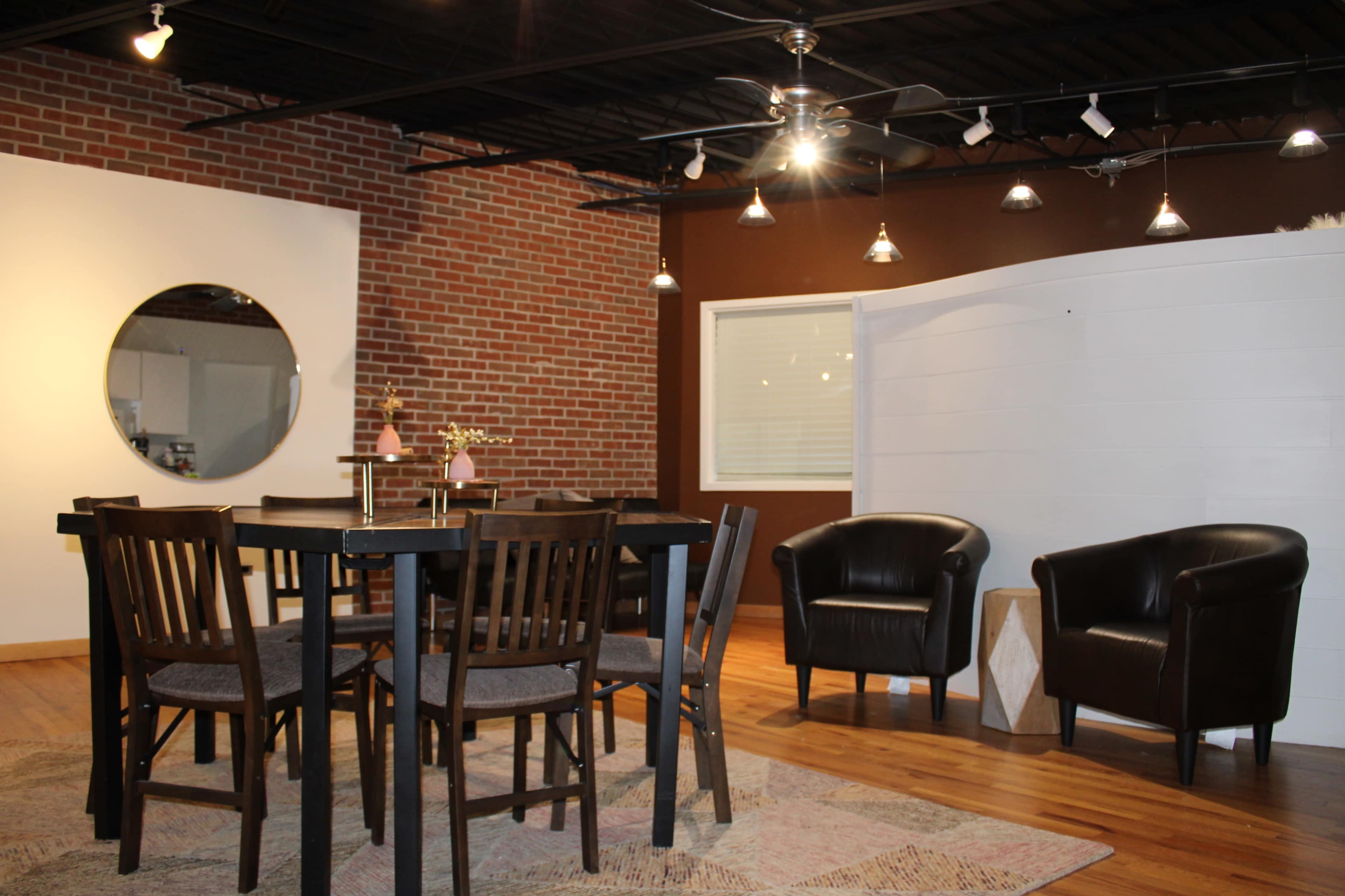 An interior space featuring a wooden dining table with chairs, two black armchairs, and a circular mirror on a brick wall.