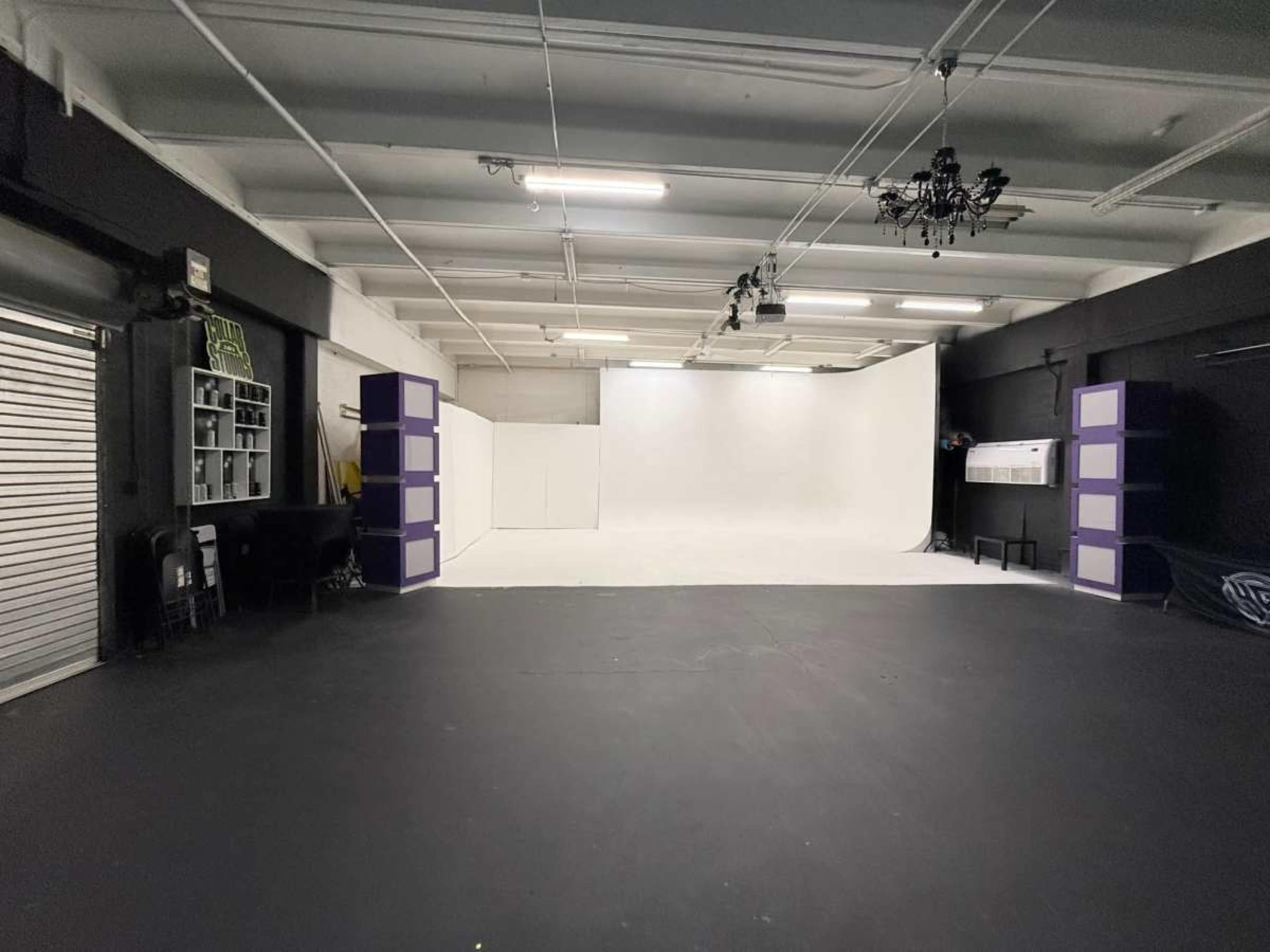 The image shows a spacious photo studio with a white backdrop, black flooring, and shelving units along the walls.