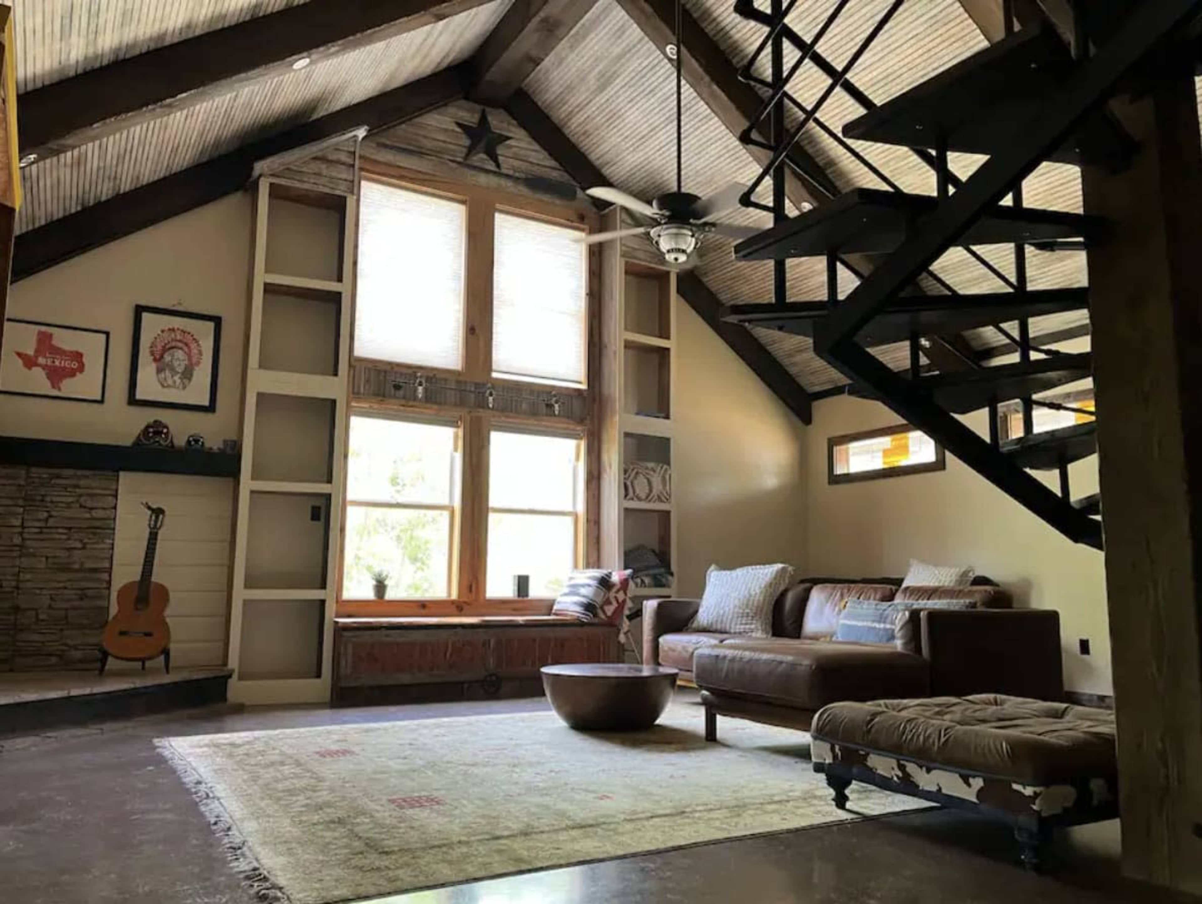 The image features a cozy living room with a wooden staircase, large windows letting in natural light, a leather sofa, and a guitar positioned near a stone accent wall.