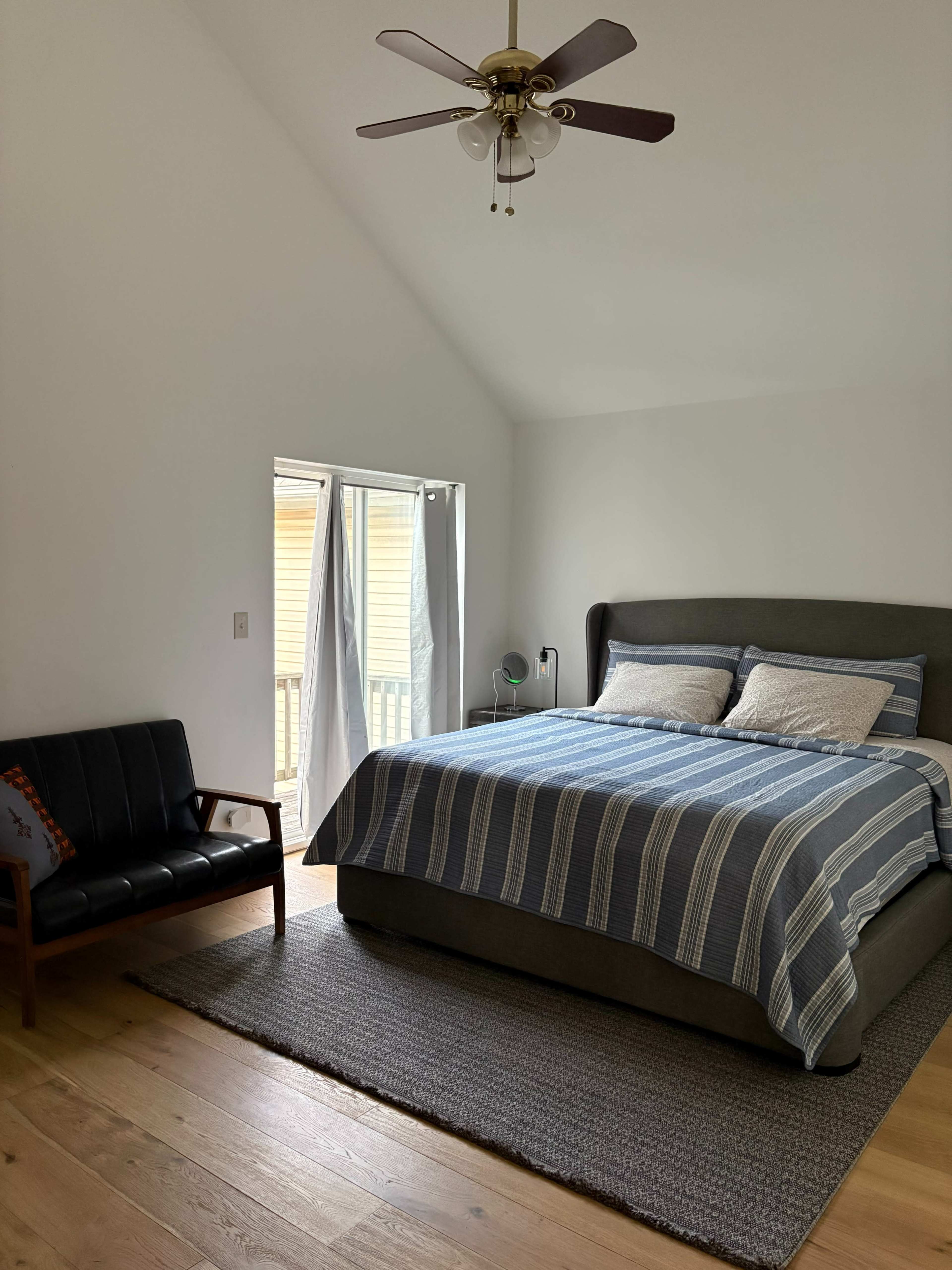 The image shows a simple bedroom featuring a large bed with a striped blue and white bedspread, a couple of pillows, a ceiling fan, a chair beside the bed, and a set of curtains covering a sliding door.