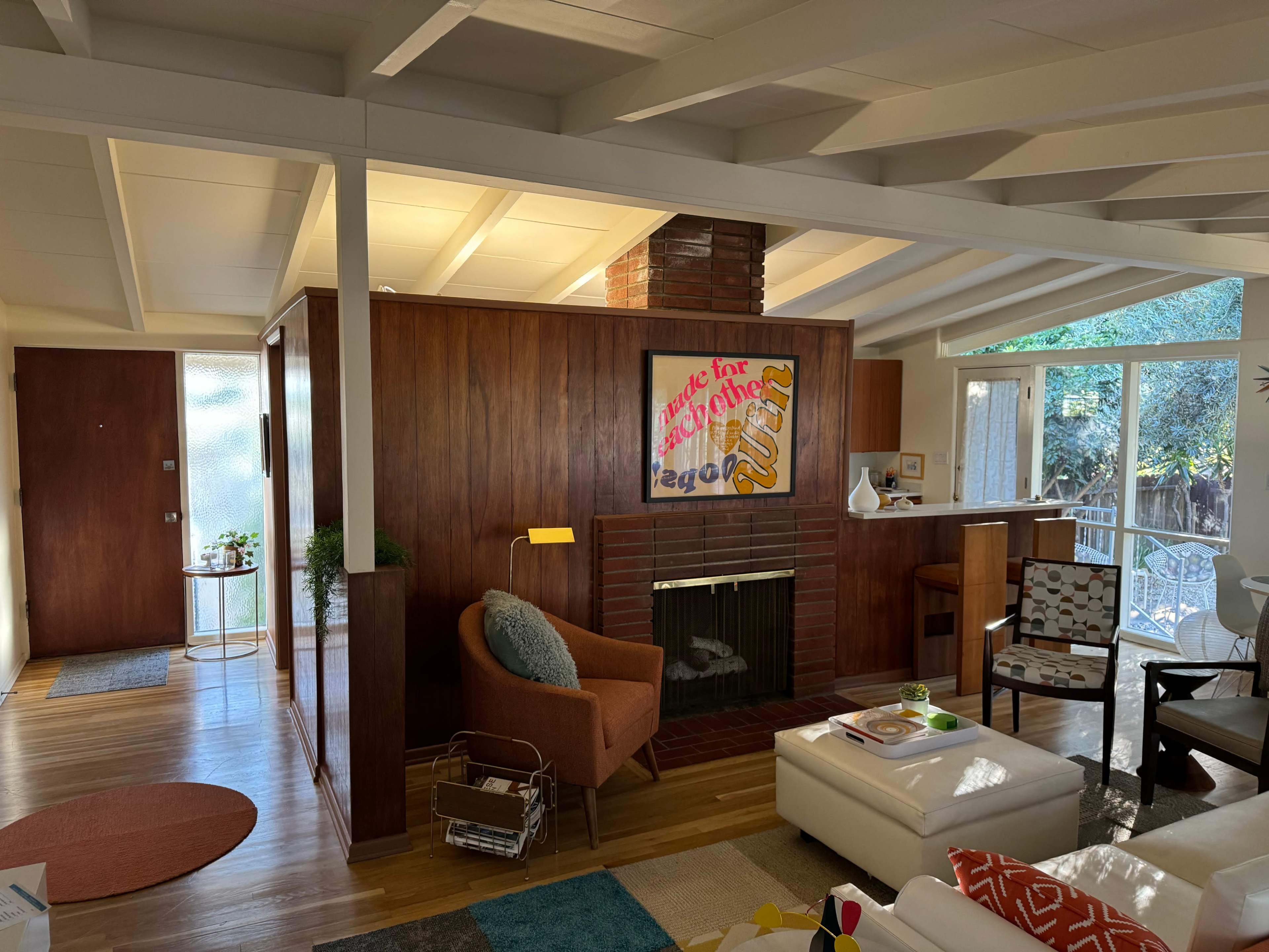 The image shows a mid-century modern living room featuring a fireplace, wooden paneling, and large windows that allow natural light to enter the space.