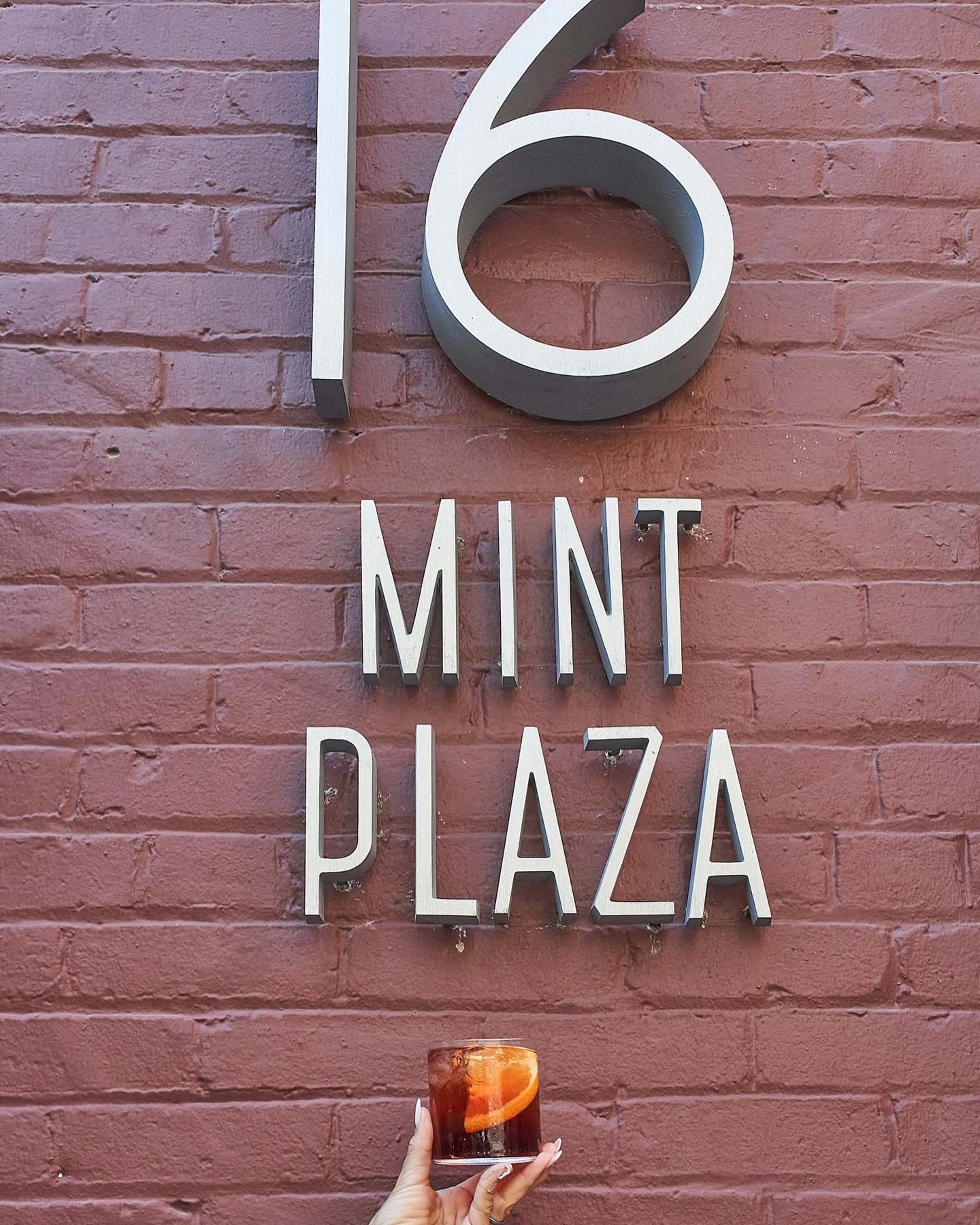 A hand holds a drink in front of a brick wall displaying the number "16" and the words "MINT PLAZA."