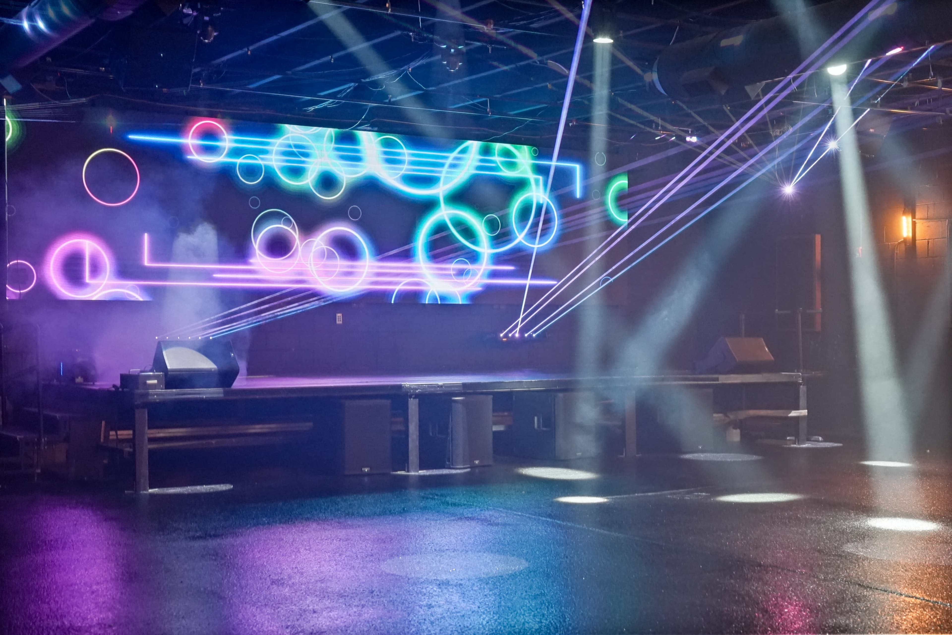 The image shows a dimly lit performance space with a stage, colored lights, and beams of light creating a vibrant atmosphere.