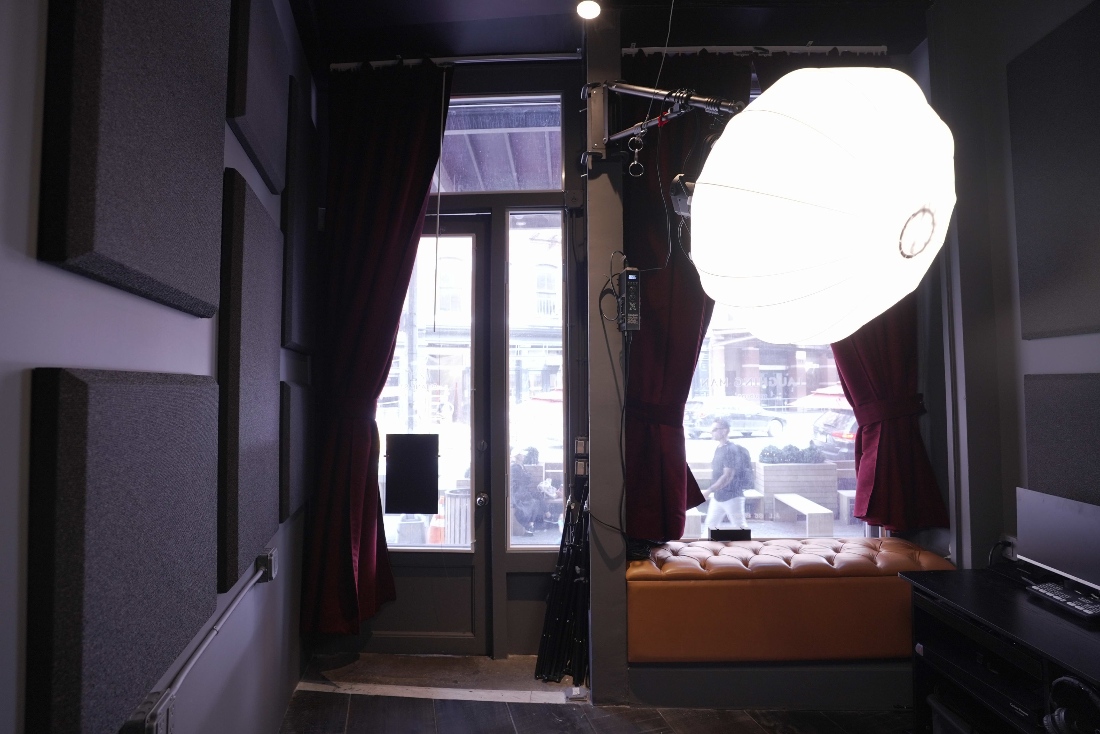 The image shows a cozy indoor space with a large round lighting fixture suspended from the ceiling, red curtains framing a window, and a brown leather bench against the wall.