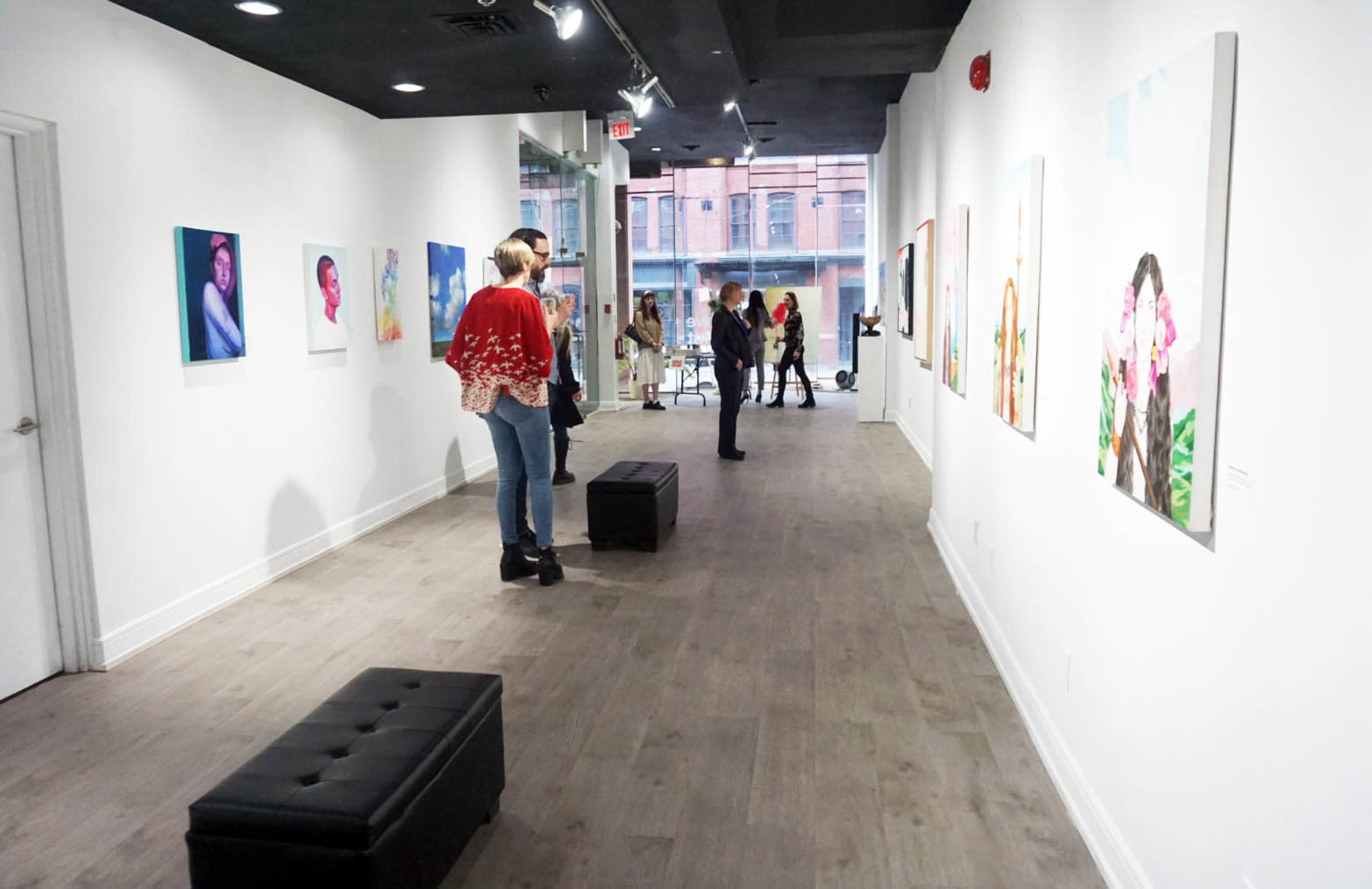 People are viewing artwork in a gallery with white walls and wooden flooring, while some benches are placed along the side.