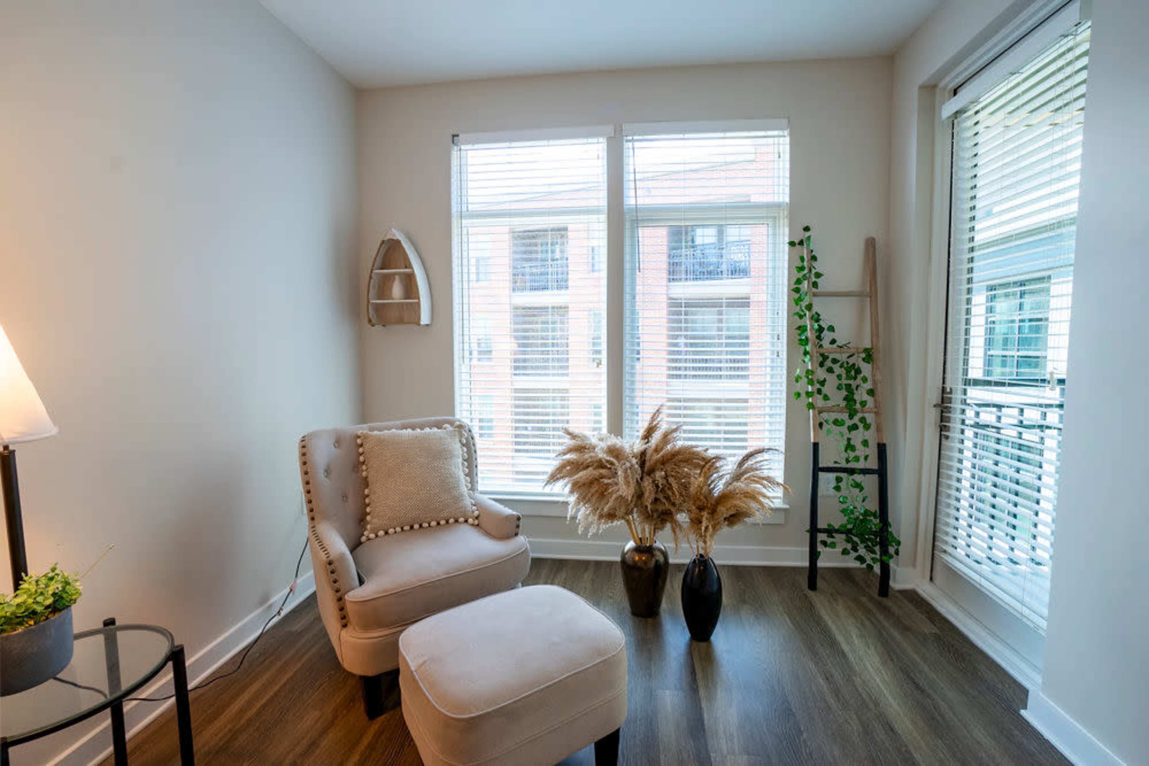 A cozy corner of a room featuring a chair and ottoman, two large windows with blinds, and decorative plants in a vase.