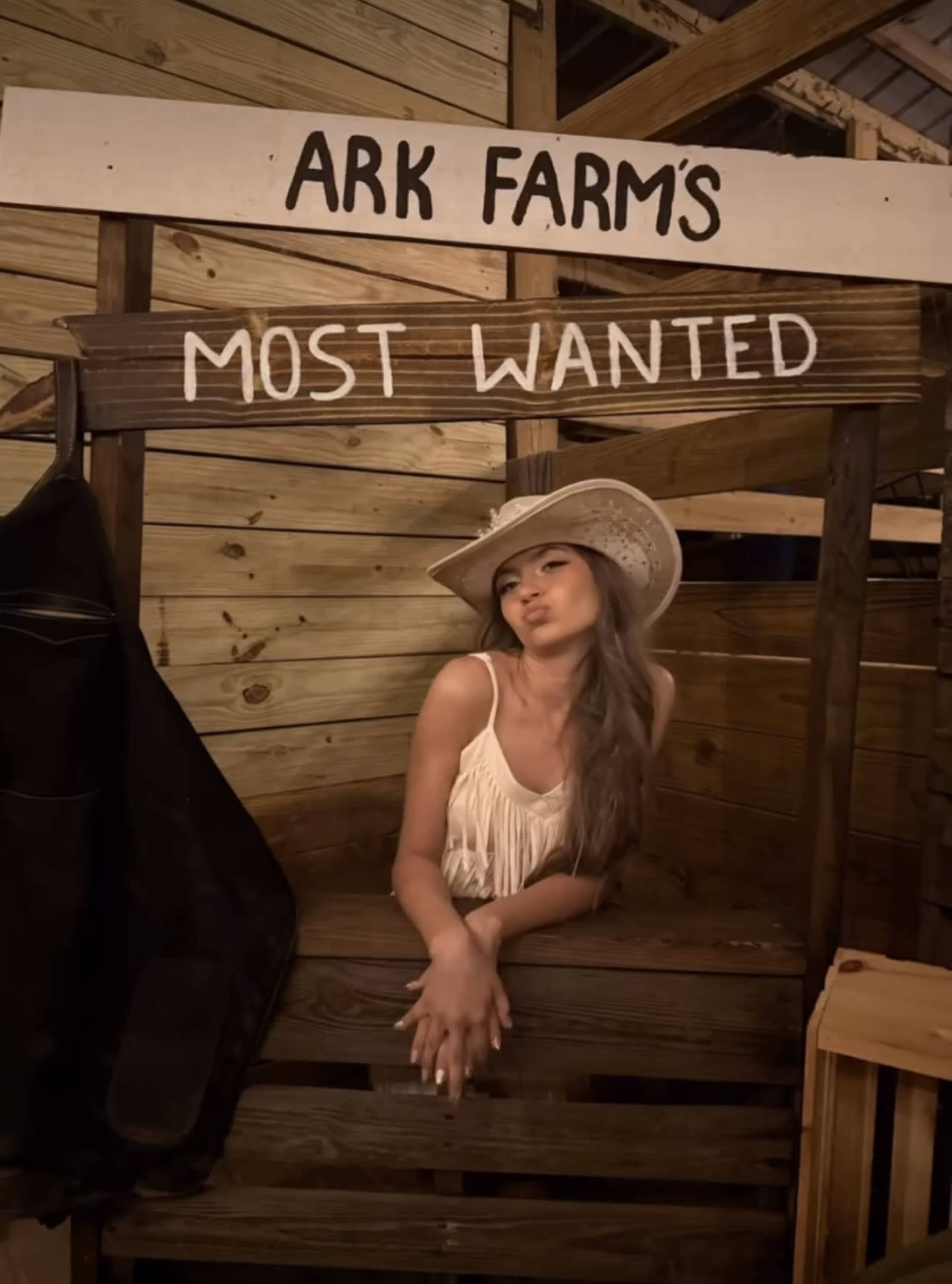 A woman in a cowboy hat poses in a wooden structure labeled "ARK FARM'S MOST WANTED."