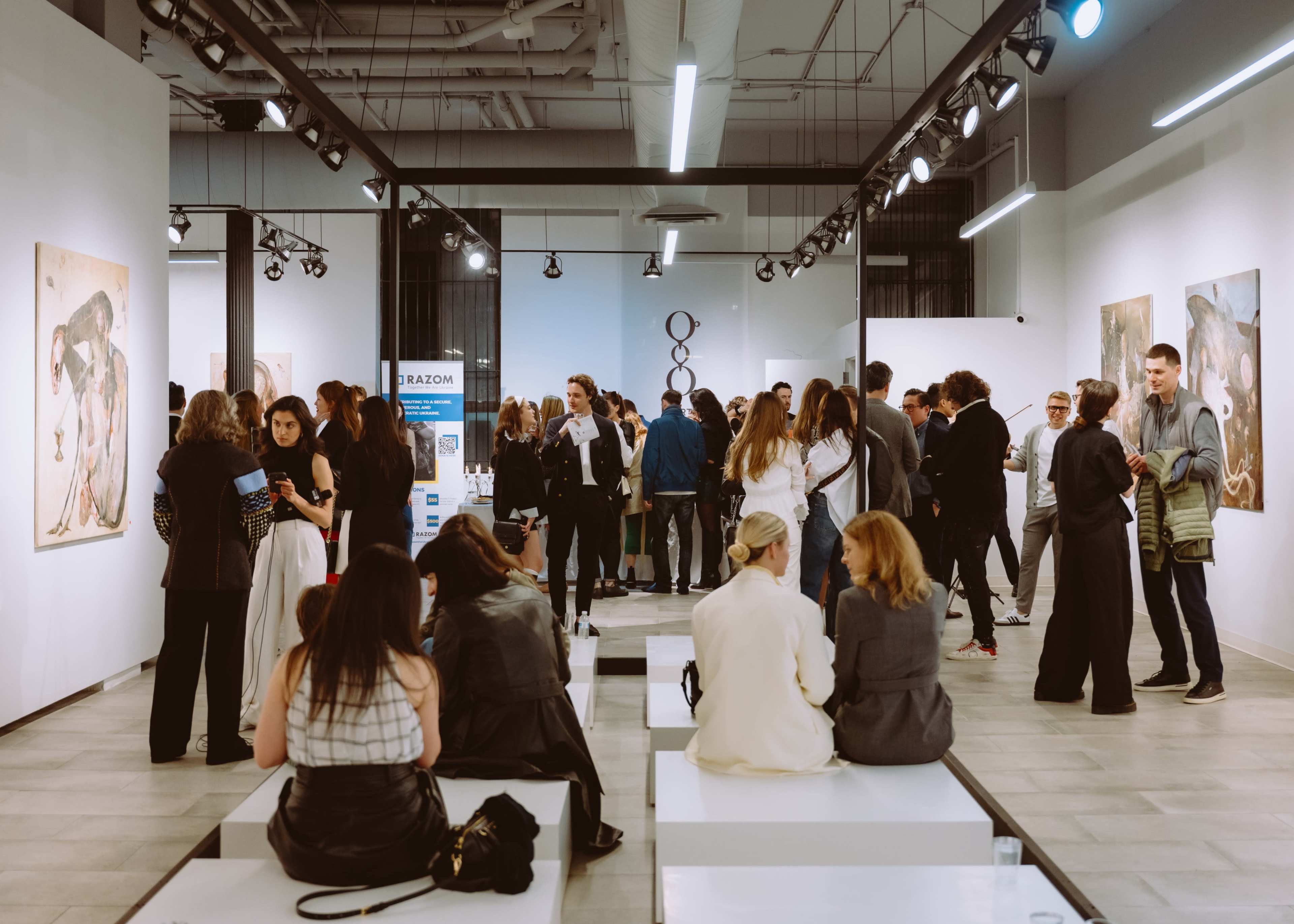 A crowded art gallery features attendees socializing in front of various paintings displayed on the walls.