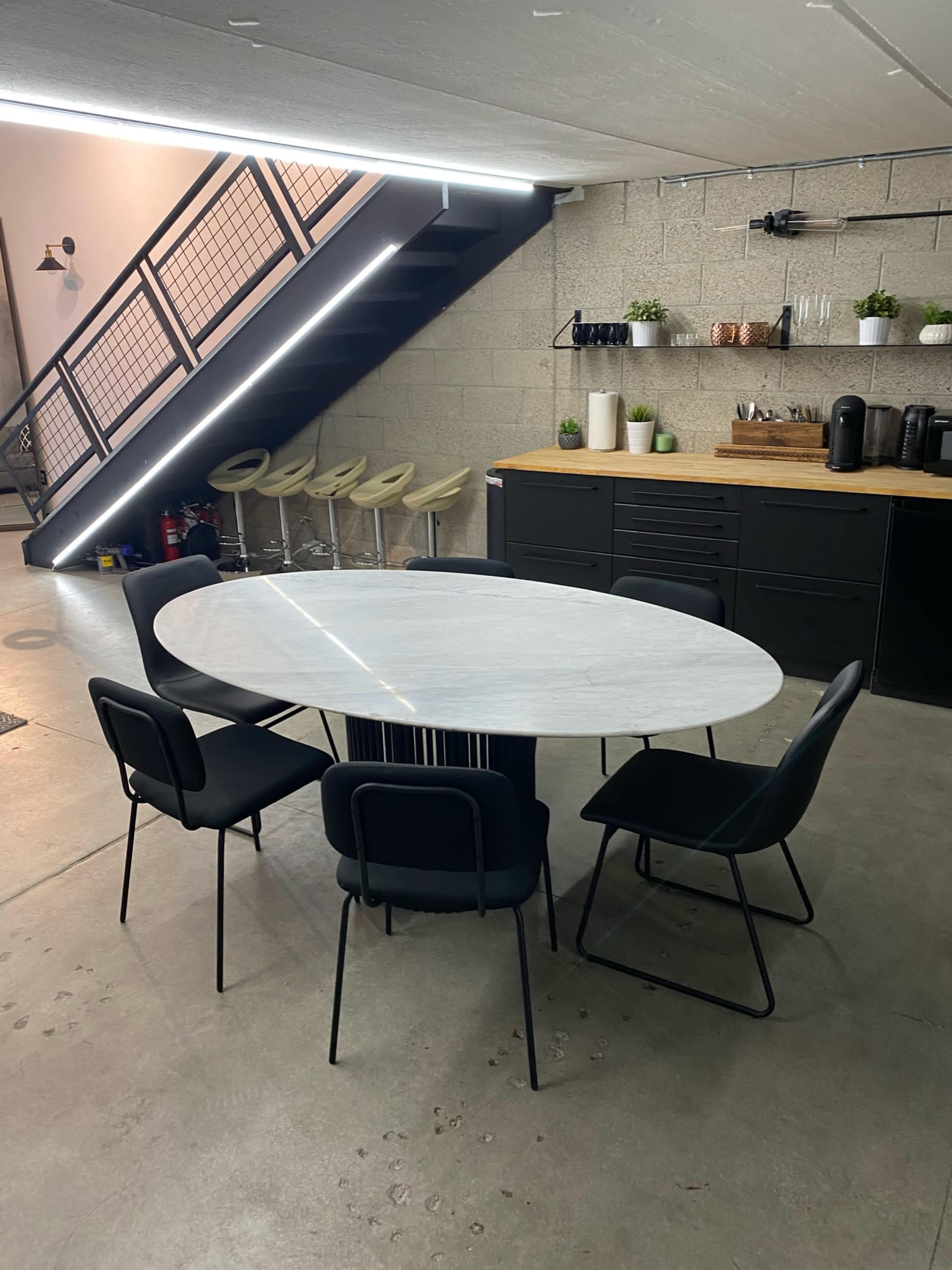 The image shows a modern dining area with a round marble table surrounded by six black chairs, located under a staircase in a minimalist space with a kitchenette in the background.