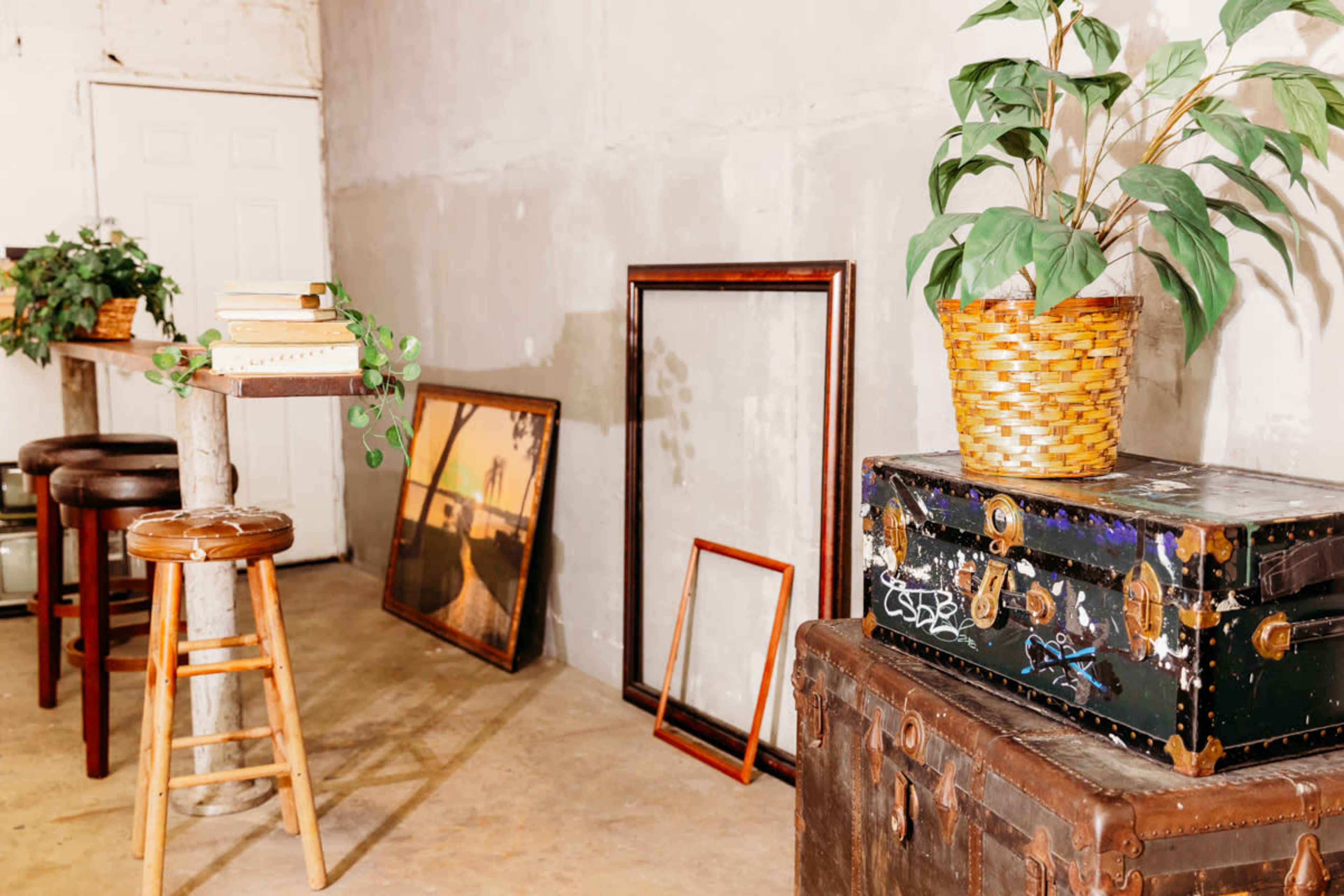 A room with bare concrete walls featuring a wooden bar, stools, a potted plant, and various frames and trunks arranged on the floor.