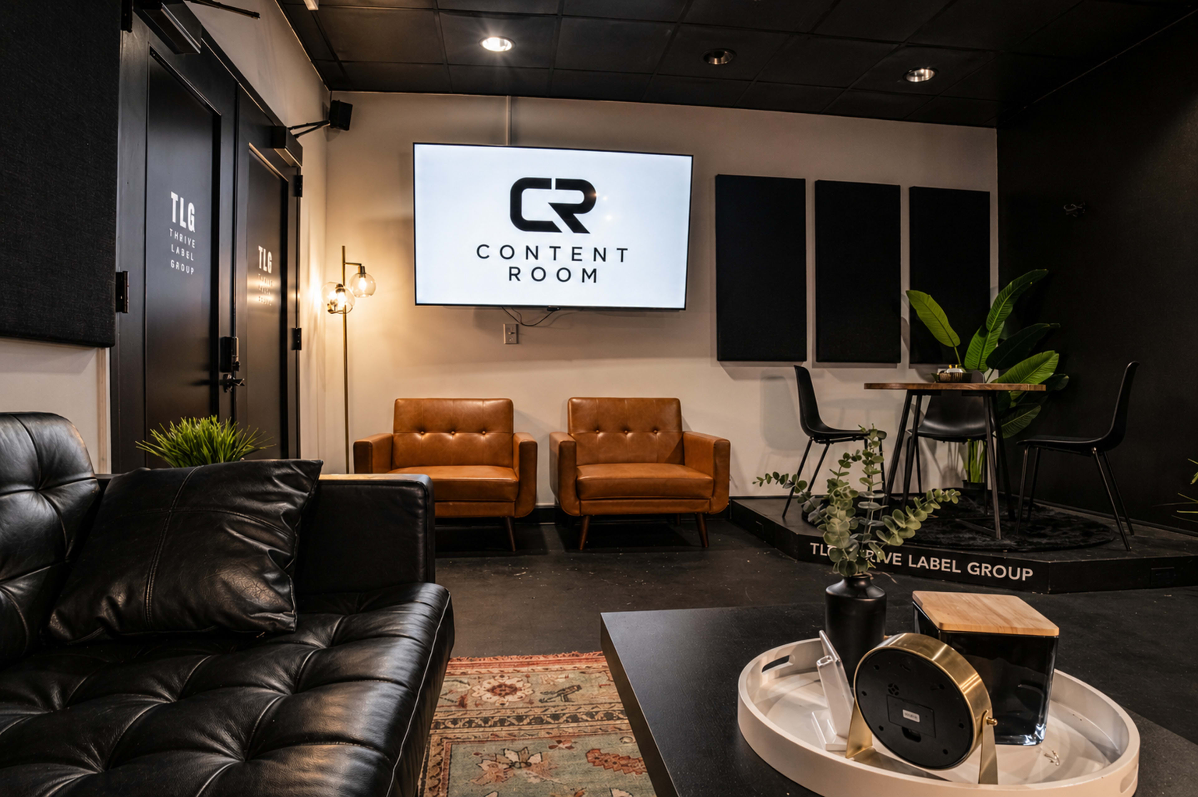 The image shows a modern content creation room featuring a large screen displaying "CONTENT ROOM," with leather chairs, a black couch, and a small table adorned with decorative plants.