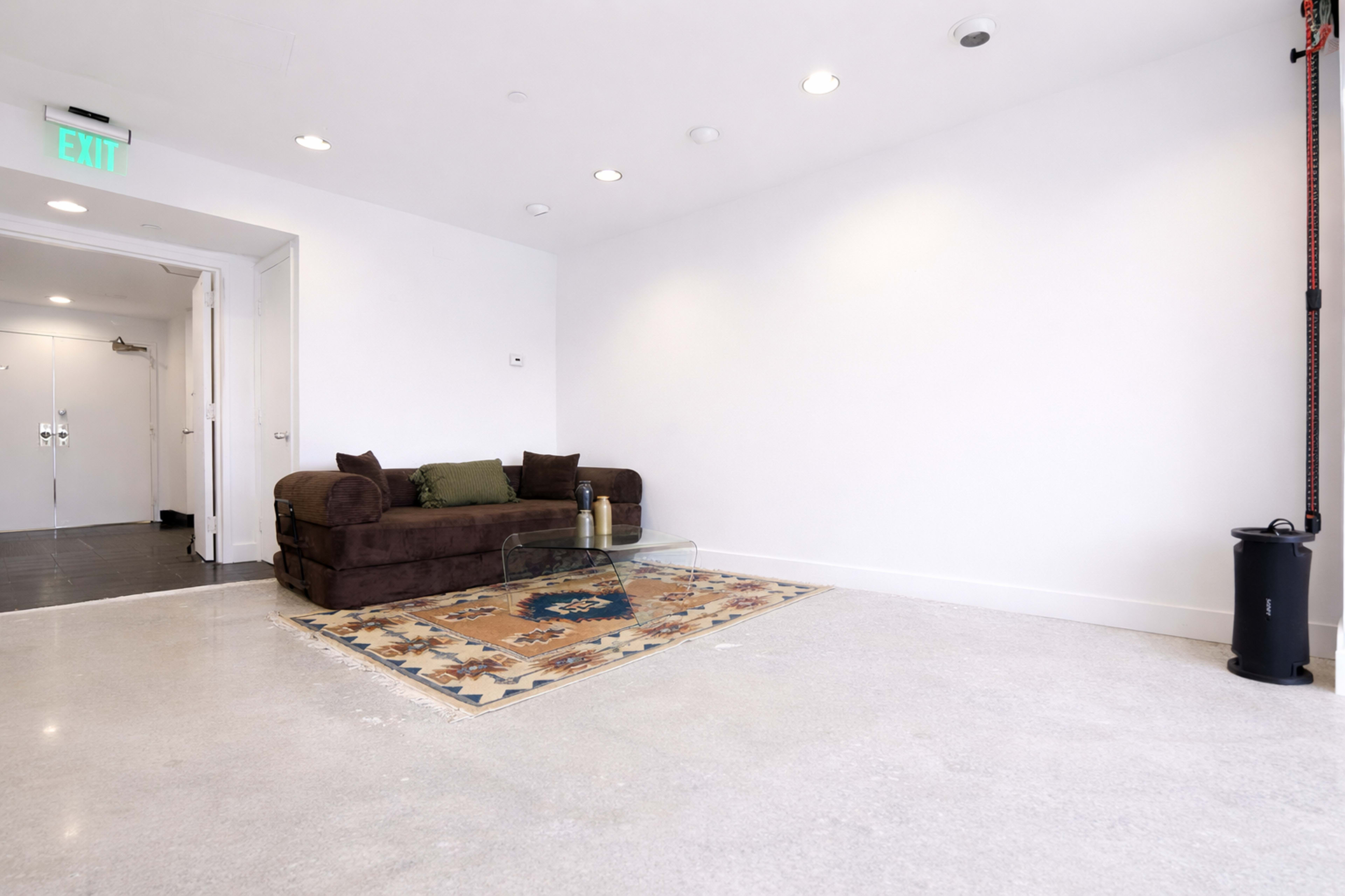 The image shows a minimalist interior with a brown couch and a glass coffee table placed on a patterned rug, against a white wall.