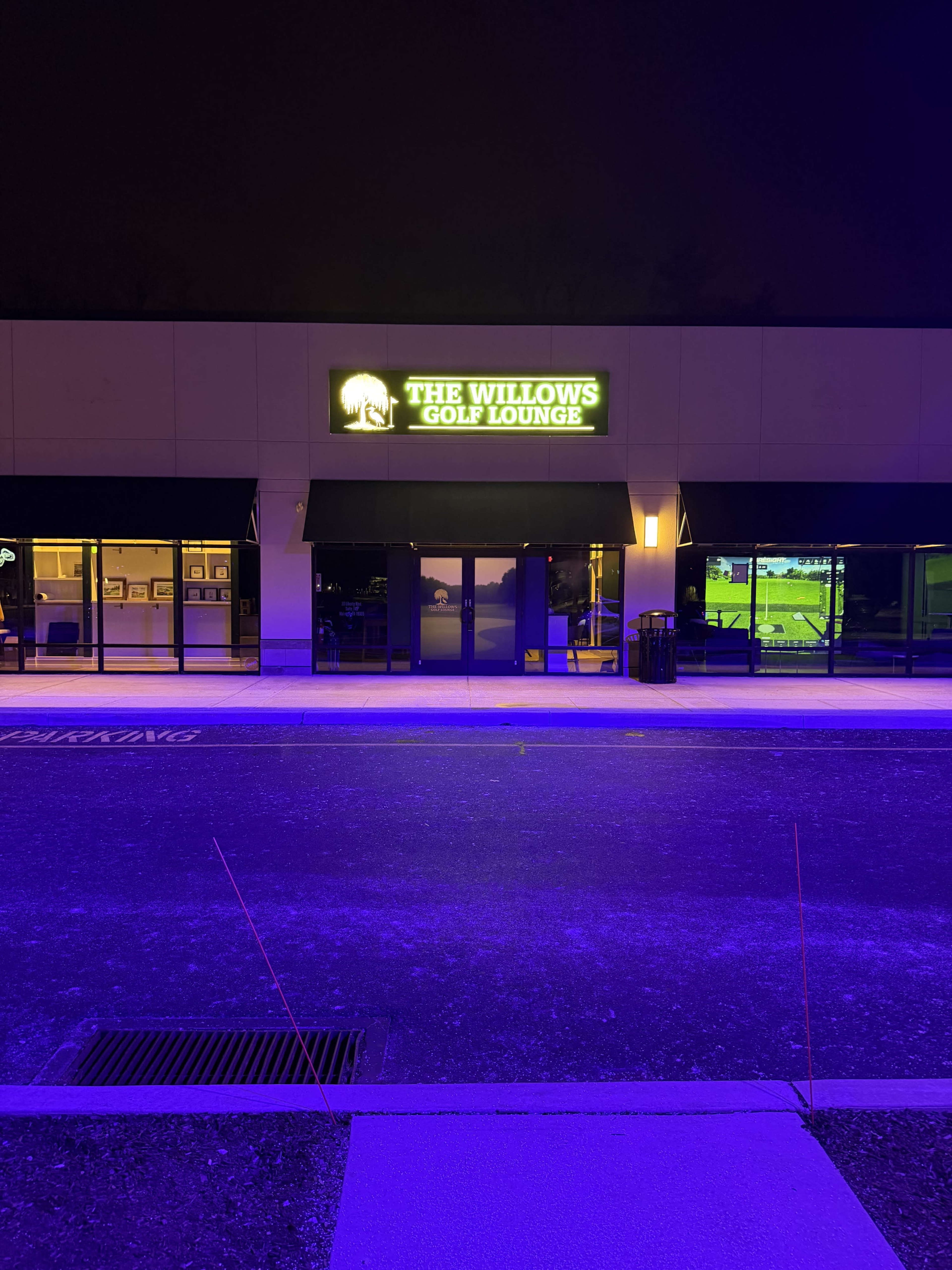 The exterior of The Willows Golf Lounge is illuminated by a bright sign in a dark setting, with a parking lot in the foreground.