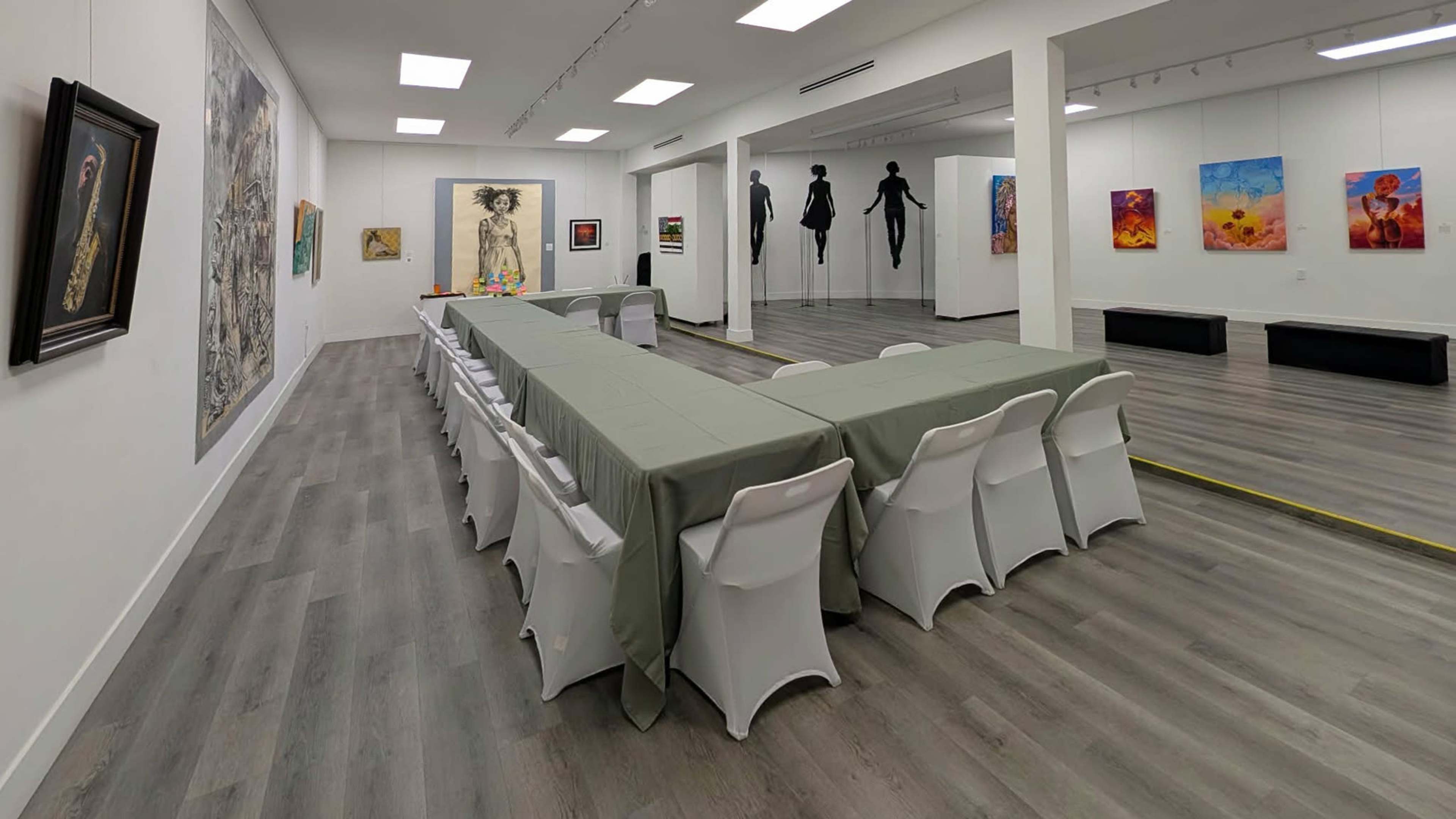 The image shows an art gallery featuring several pieces of artwork on the walls, with a long table arranged in a U-shape in the center, covered with green tablecloths and accompanied by white chairs.