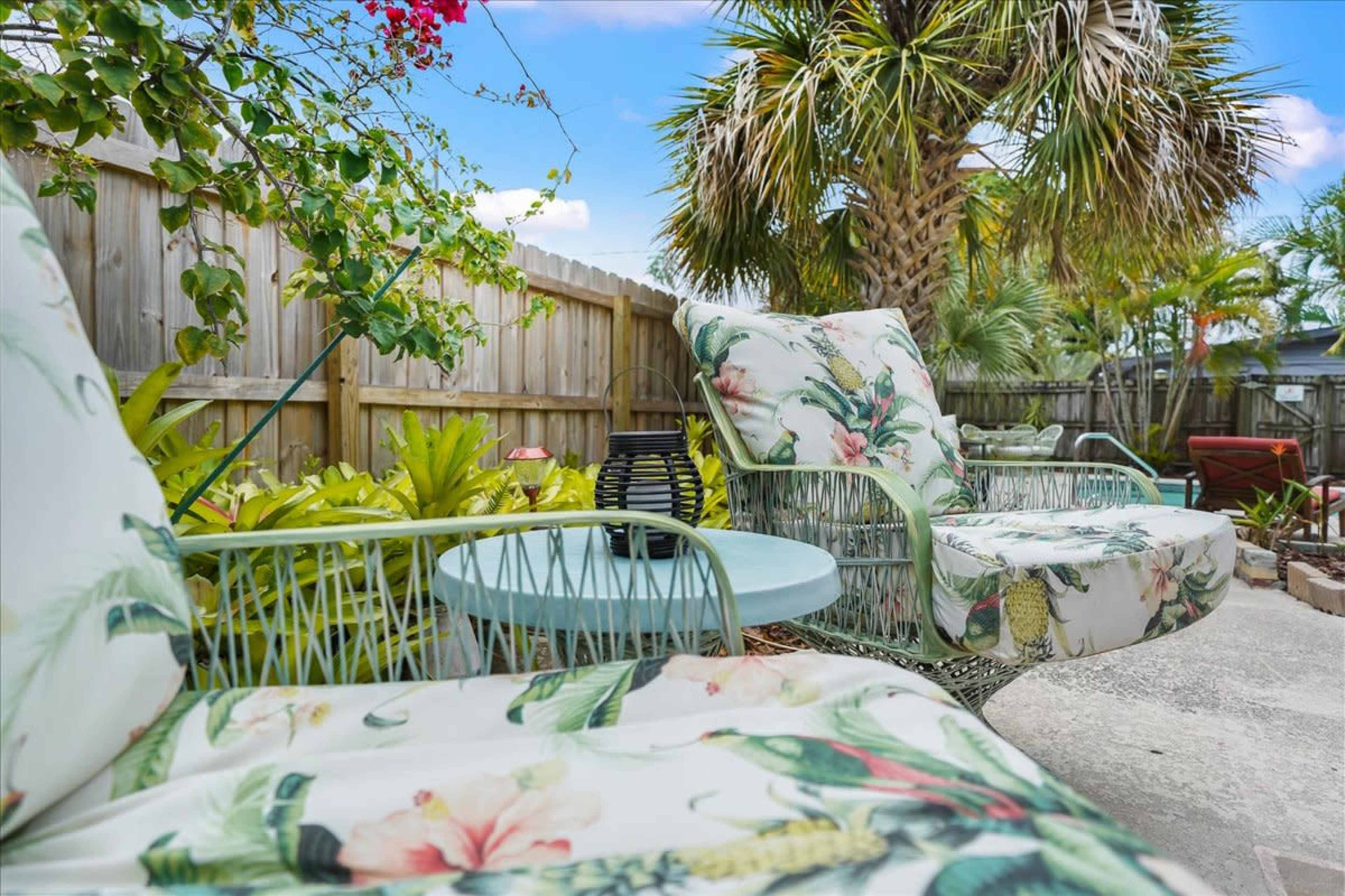 A pair of patterned outdoor chairs sit beside a small blue table in a lush backyard with palm trees and vibrant foliage.