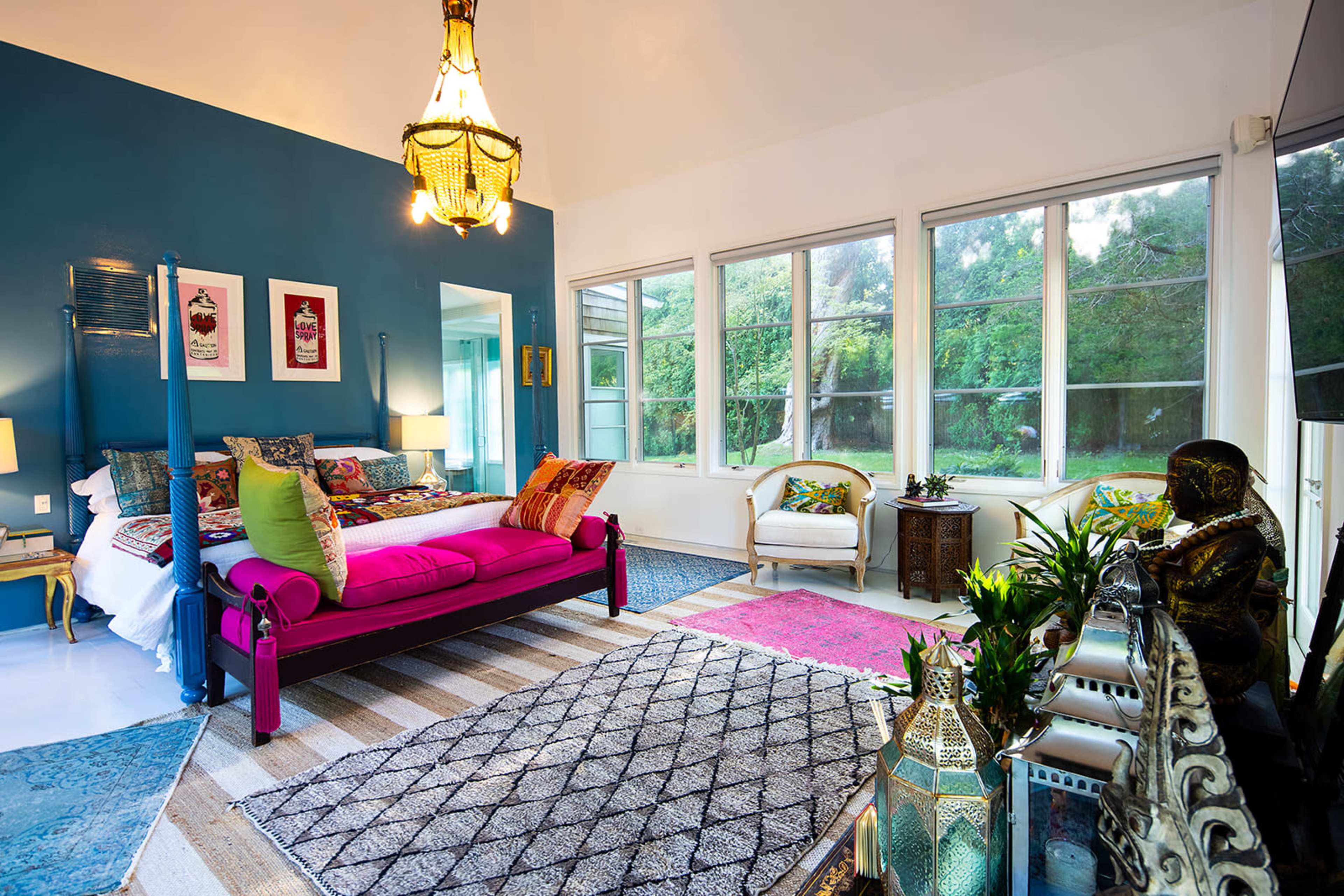 A brightly colored bedroom features a pink bed, large windows with greenery outside, and a mix of decorative furnishings.