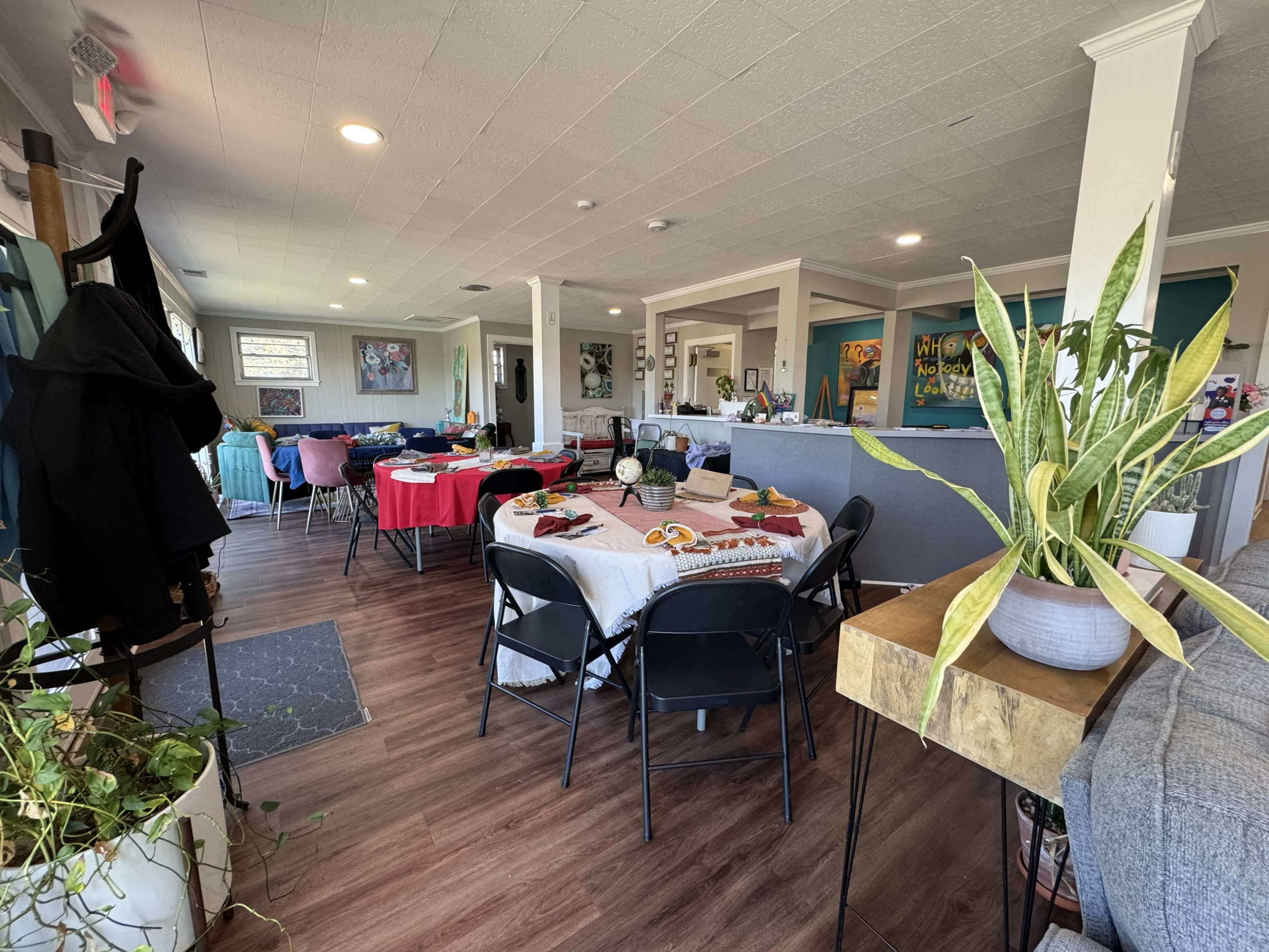 The image shows a spacious room set up for dining, featuring several tables with various colored tablecloths and decorative plants in the foreground.