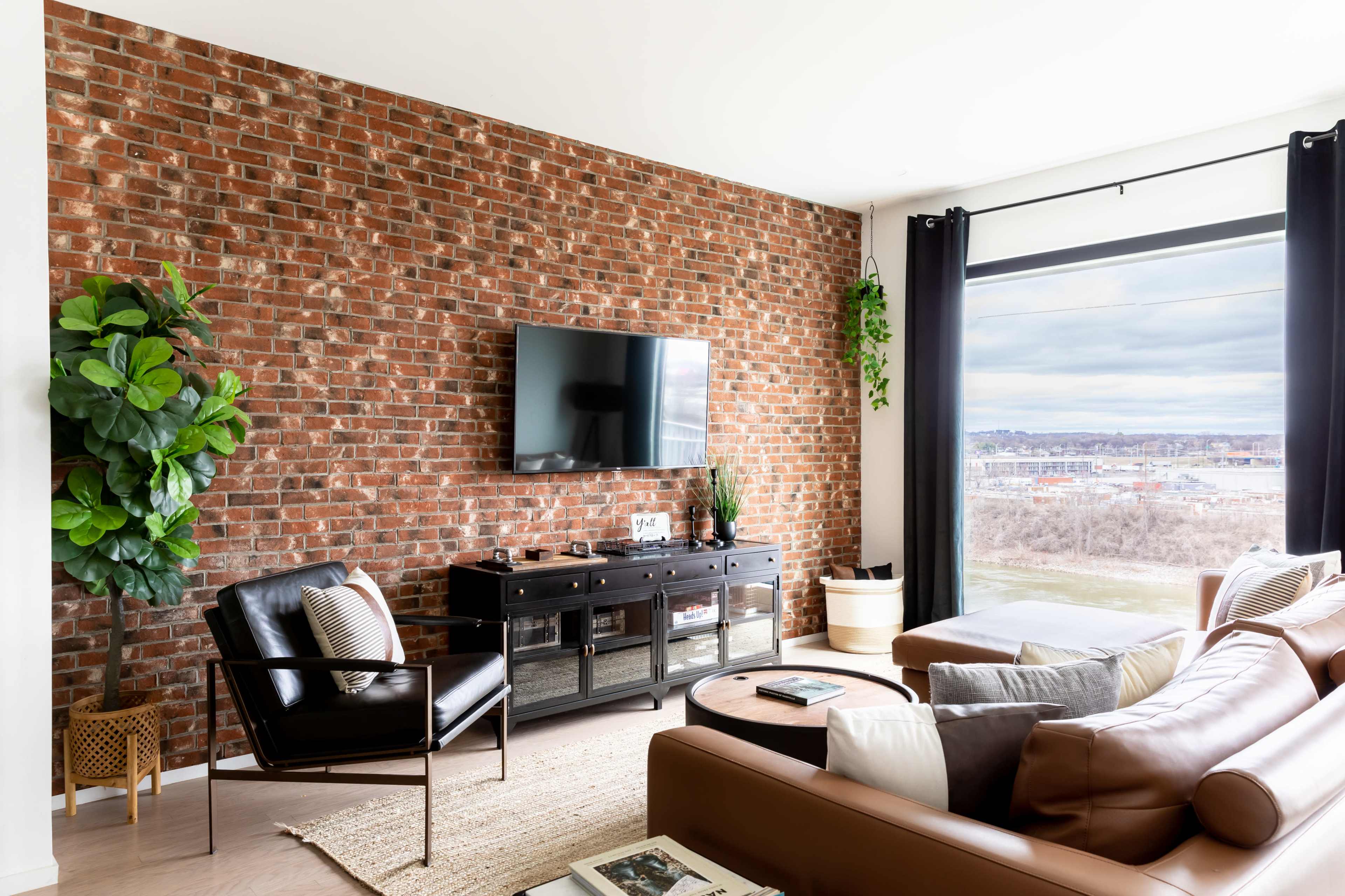 A modern living room features a brick accent wall, a large TV mounted above a dark sideboard, and a sectional sofa near a window with a view.