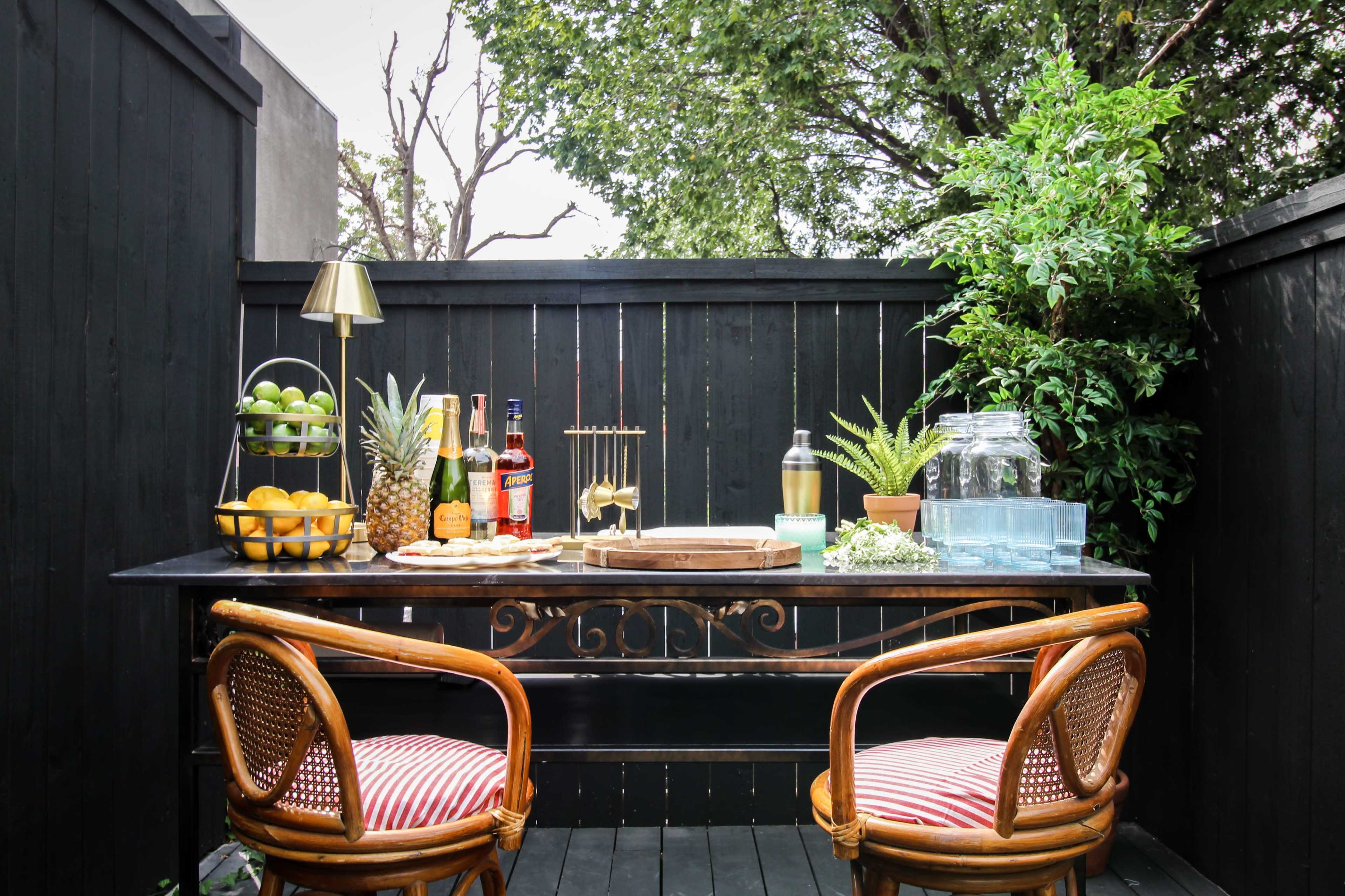 A well-set outdoor bar table adorned with drinks, fruits, and decorative plants is positioned against a black fence.
