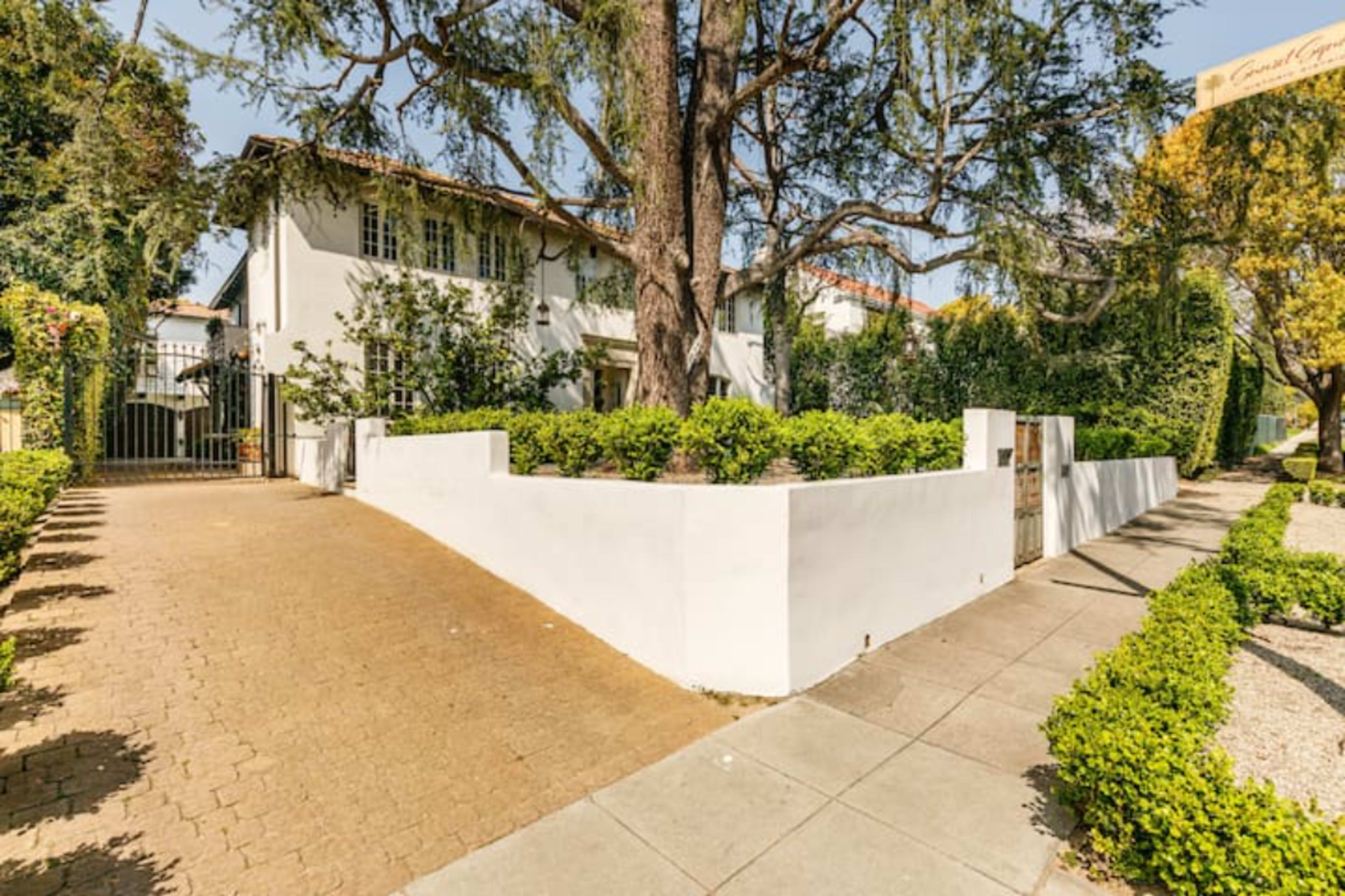 A white stucco wall encloses a landscaped garden leading to a two-story building with a tile roof, situated along a pathway lined with hedges.