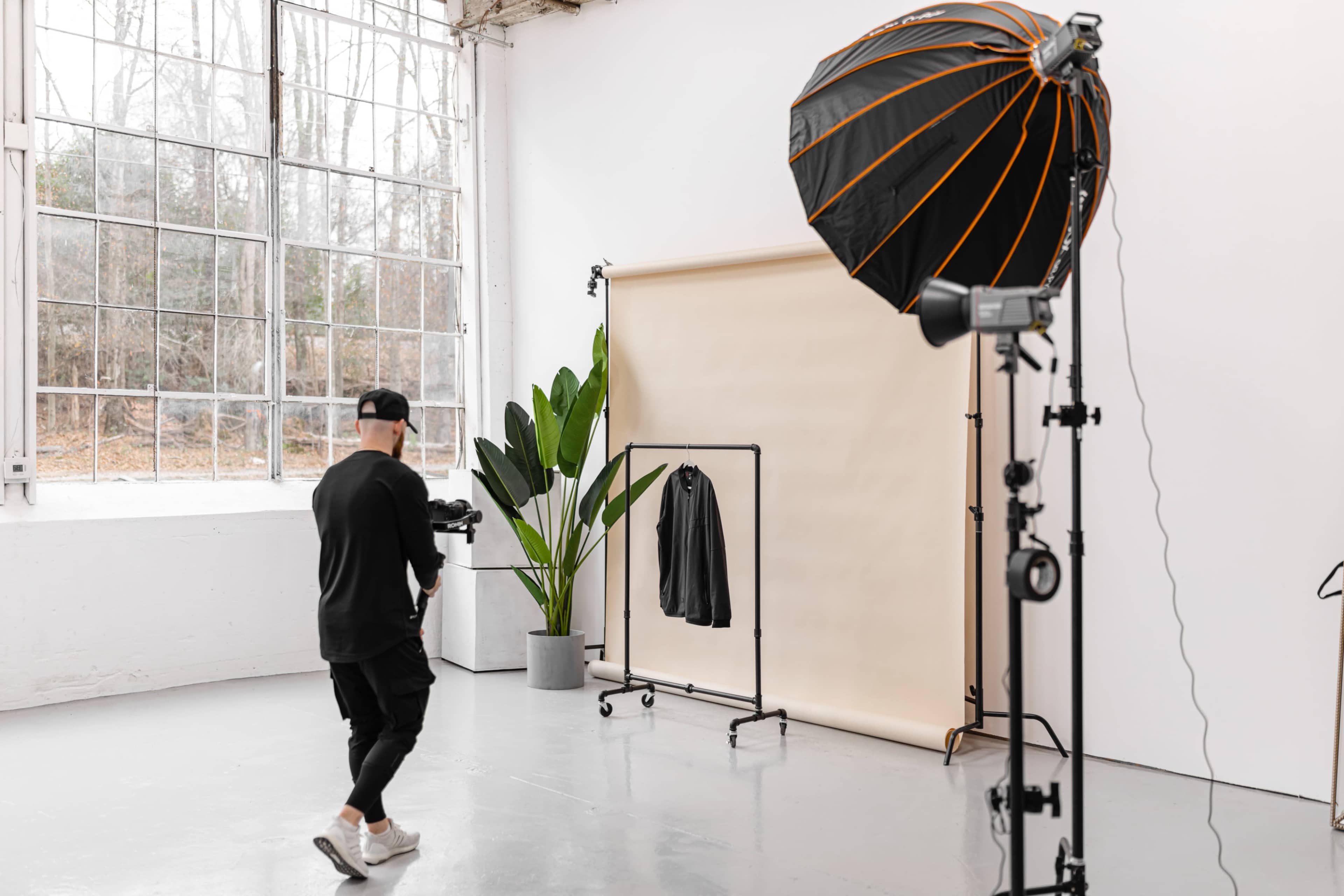 A photographer stands in a studio with a backdrop and a clothing rack holding a jacket, accompanied by a large softbox light and a potted plant.