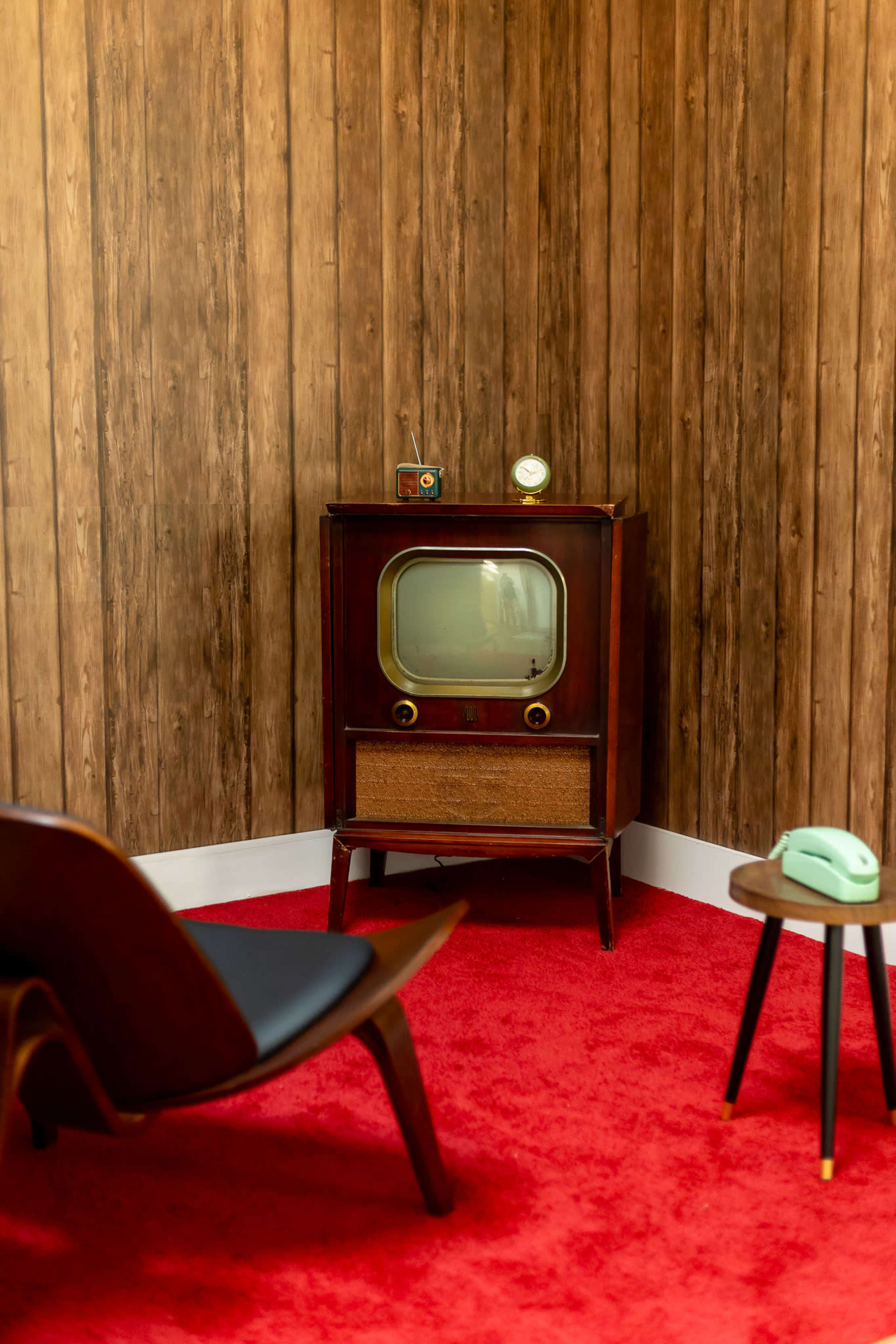 The image shows a vintage television set on a wooden floor, positioned against a wall with a wood panel finish, accompanied by a mid-century modern chair, a small side table, and a retro telephone.