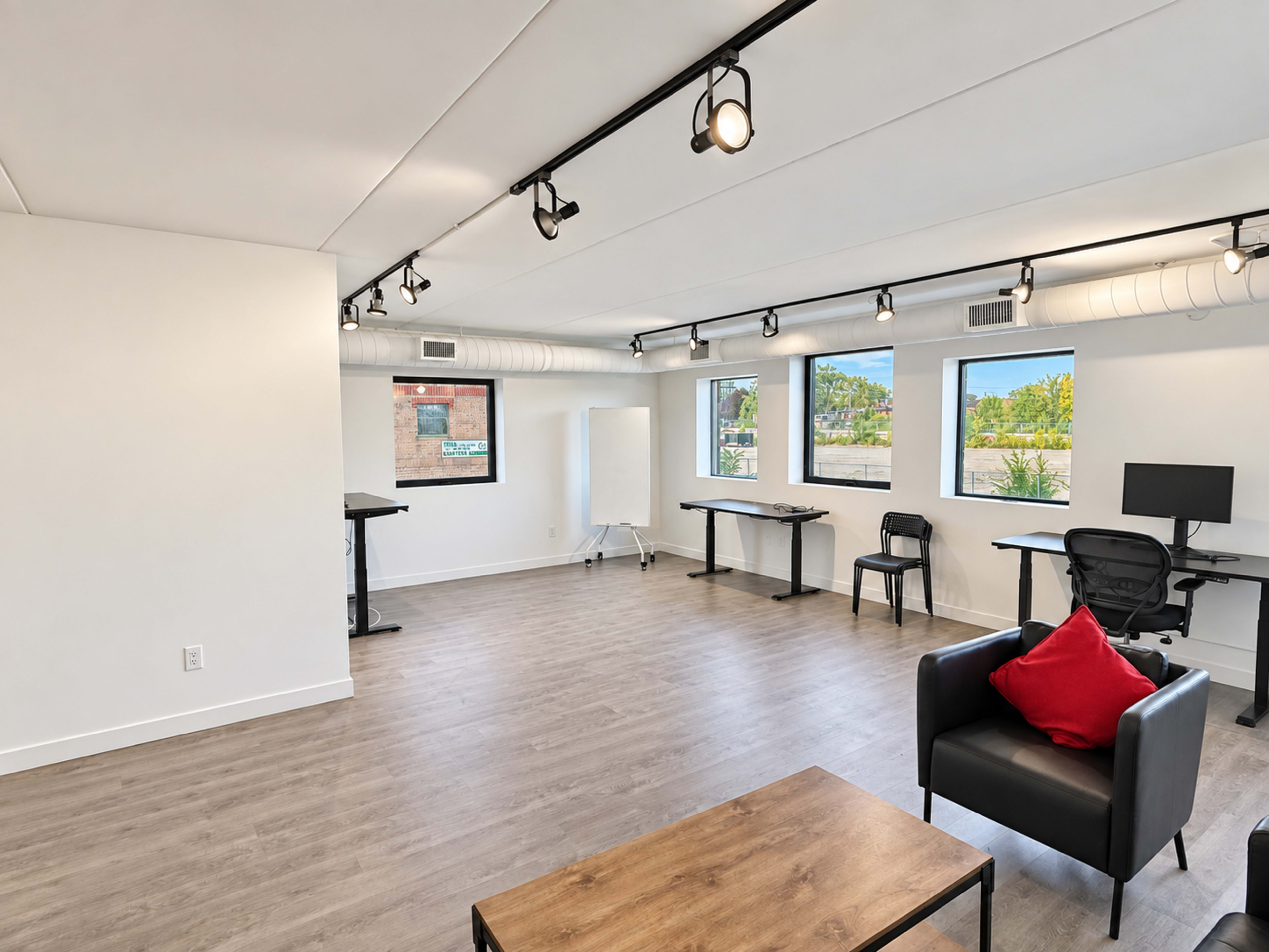The image shows a modern, sparsely furnished office space with wooden flooring, several desks, and large windows allowing natural light.