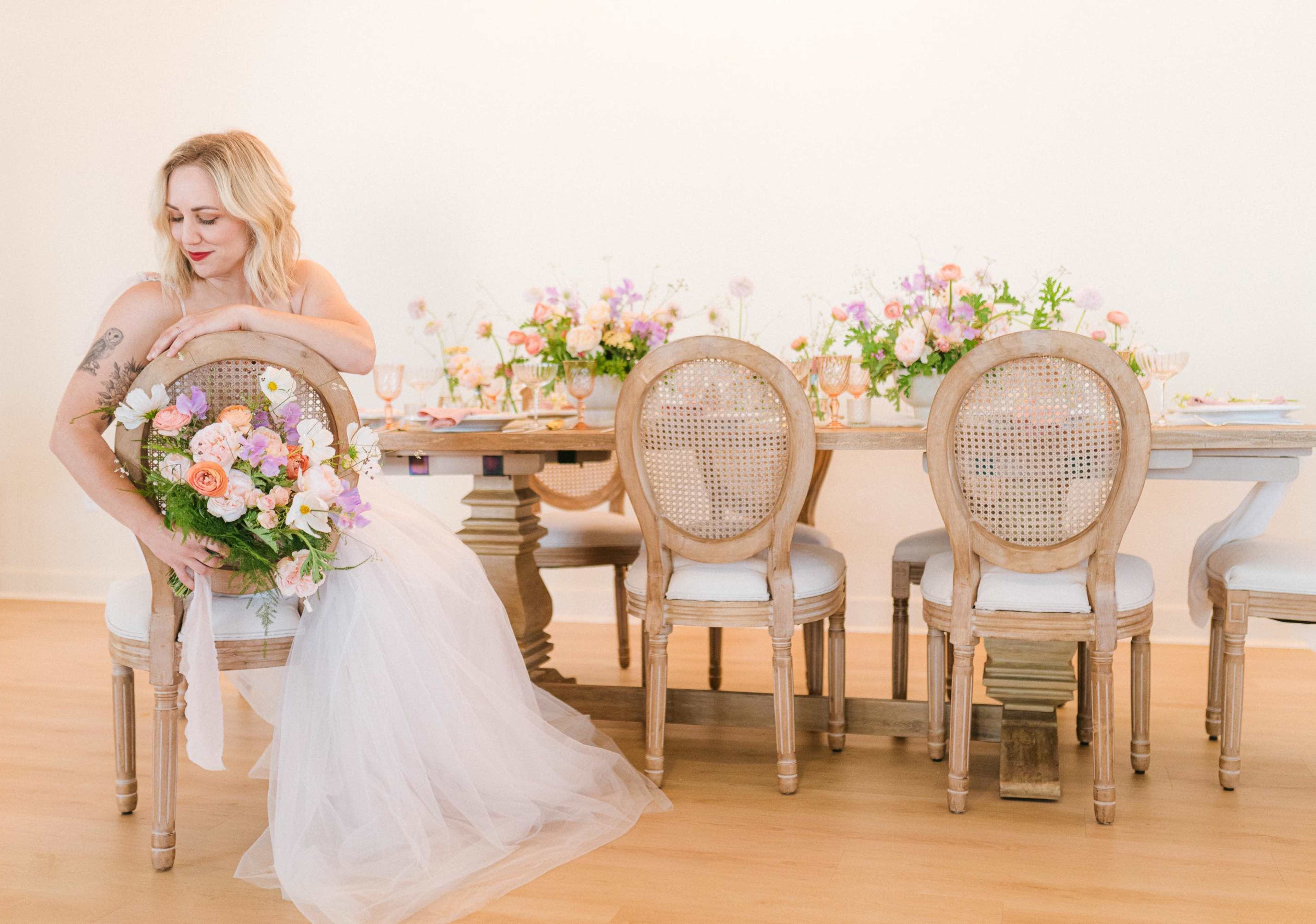 A woman in a wedding dress poses with a bouquet beside a elegantly set dining table adorned with flowers and decorative tableware.