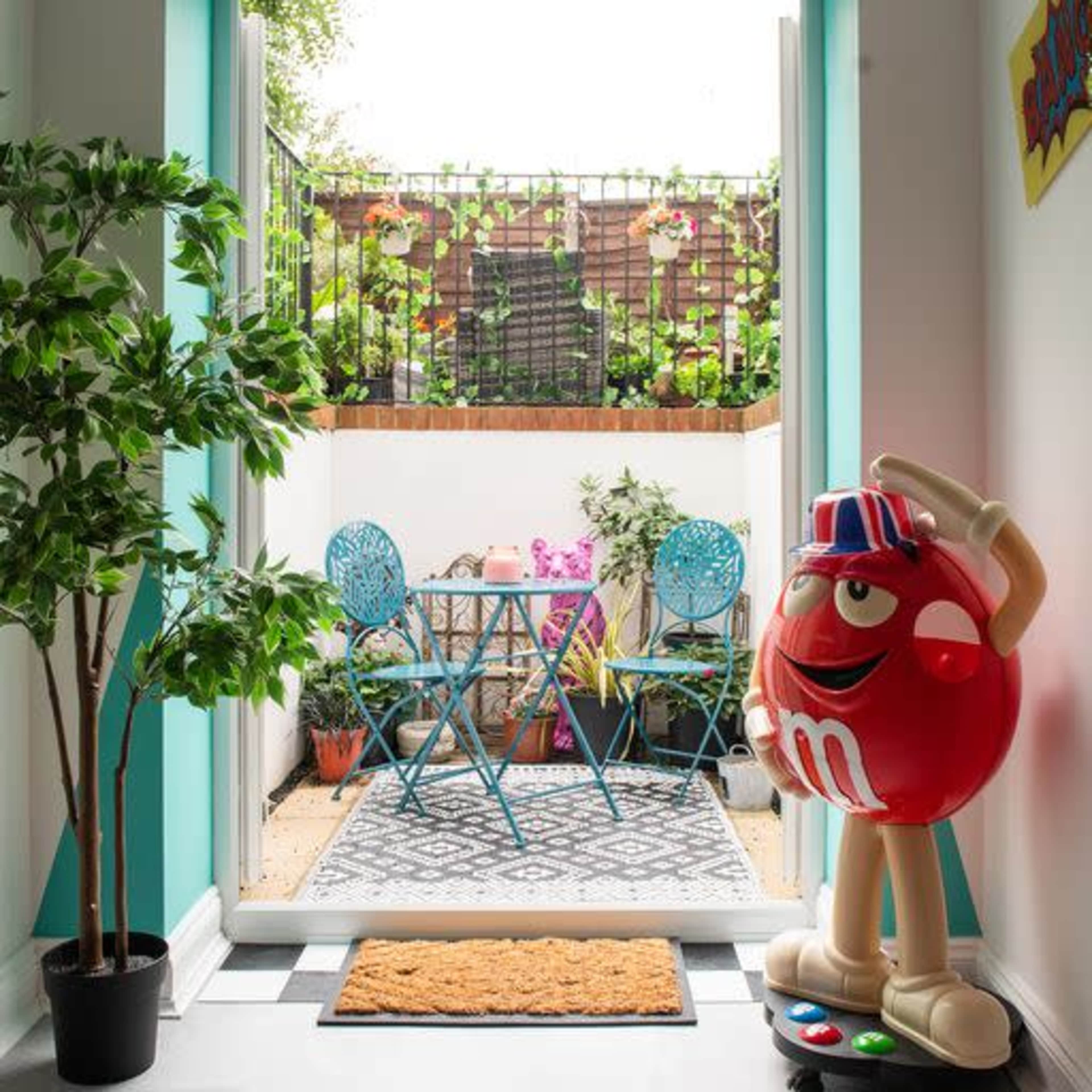 A small balcony is visible through an open door, featuring a bistro table and chairs surrounded by plants and decorative elements.