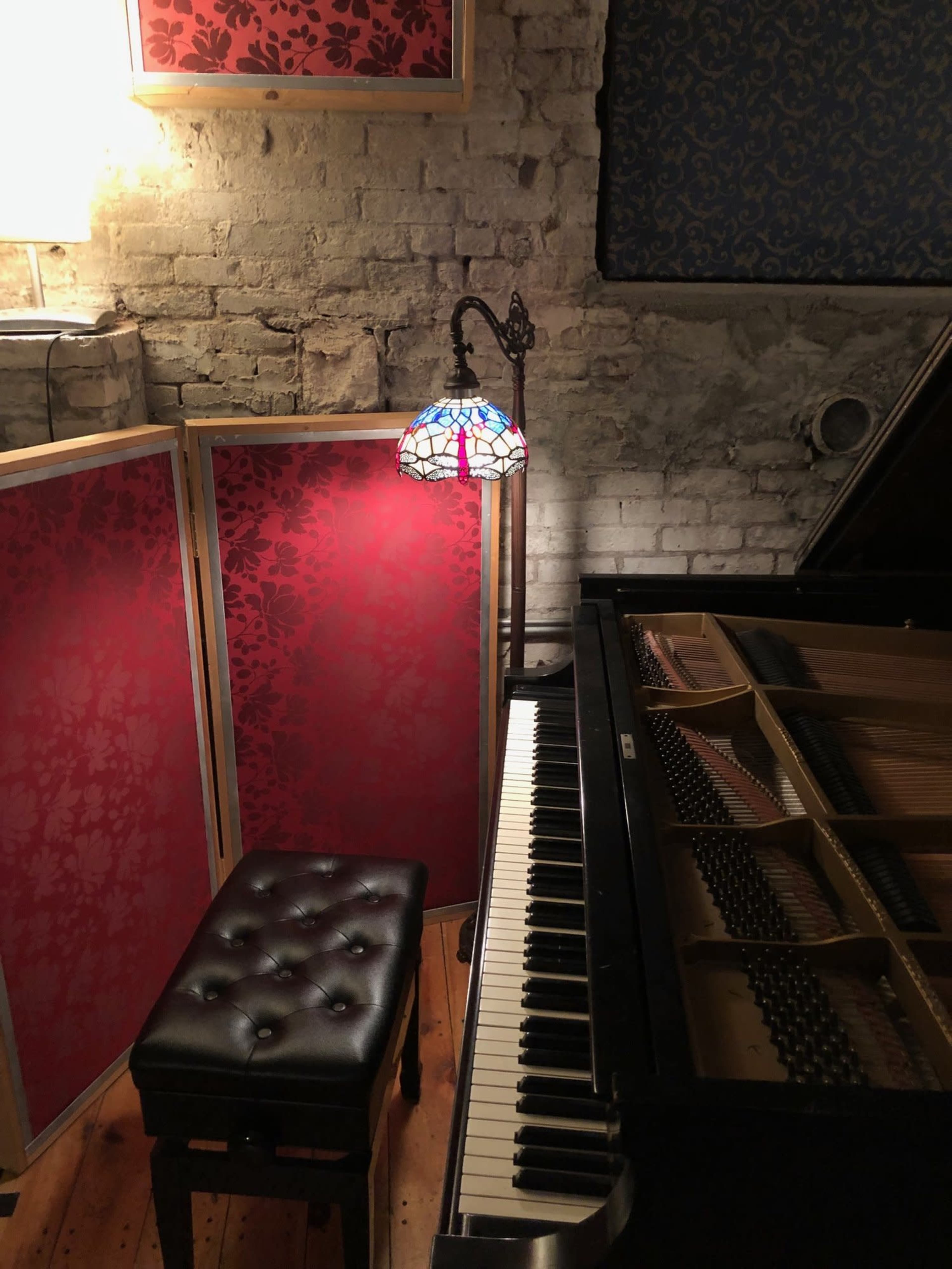 A piano with an open lid sits next to a tufted bench and a stained glass lamp, against a backdrop of patterned red screens and exposed brick walls.
