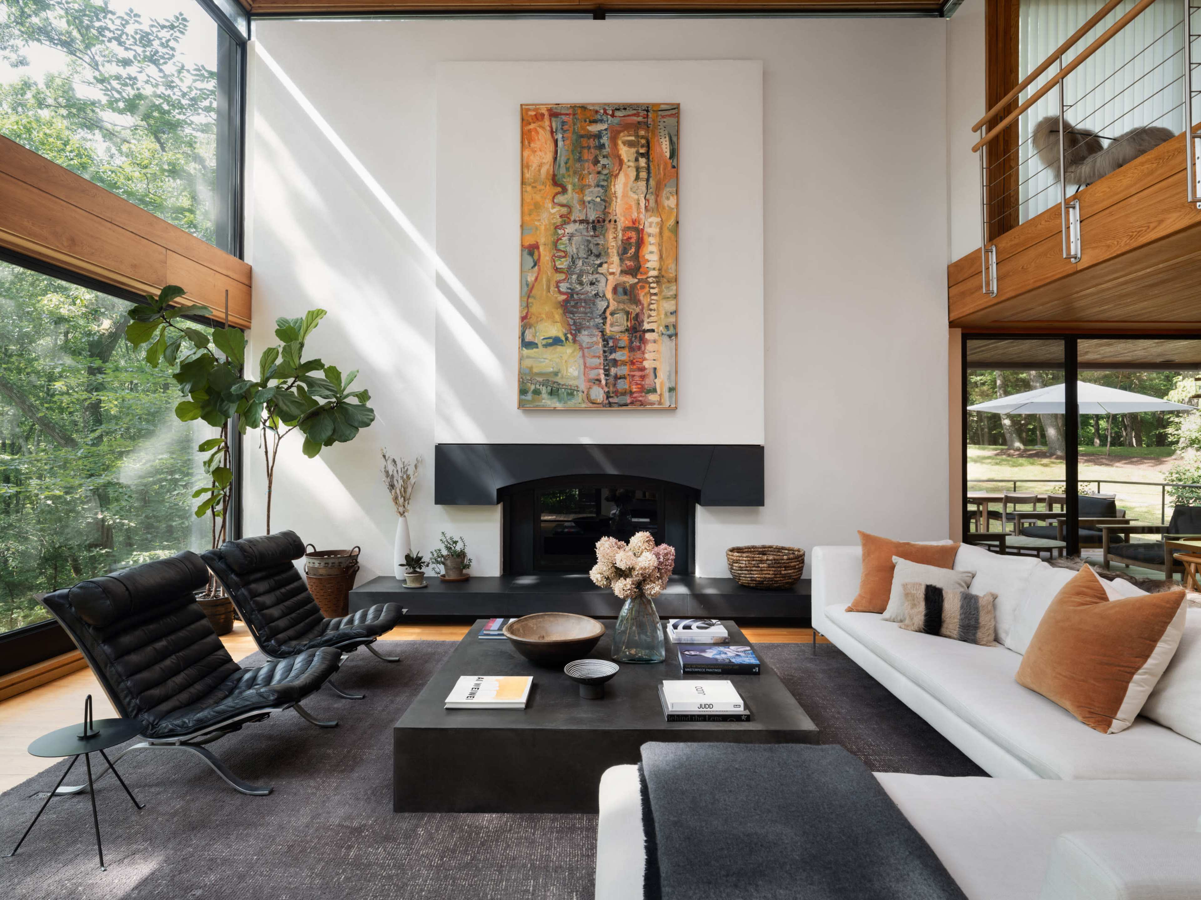The image shows a modern living room with a fireplace, large windows, and a mix of seating, including two black chairs and a white sofa, surrounded by plants and art.