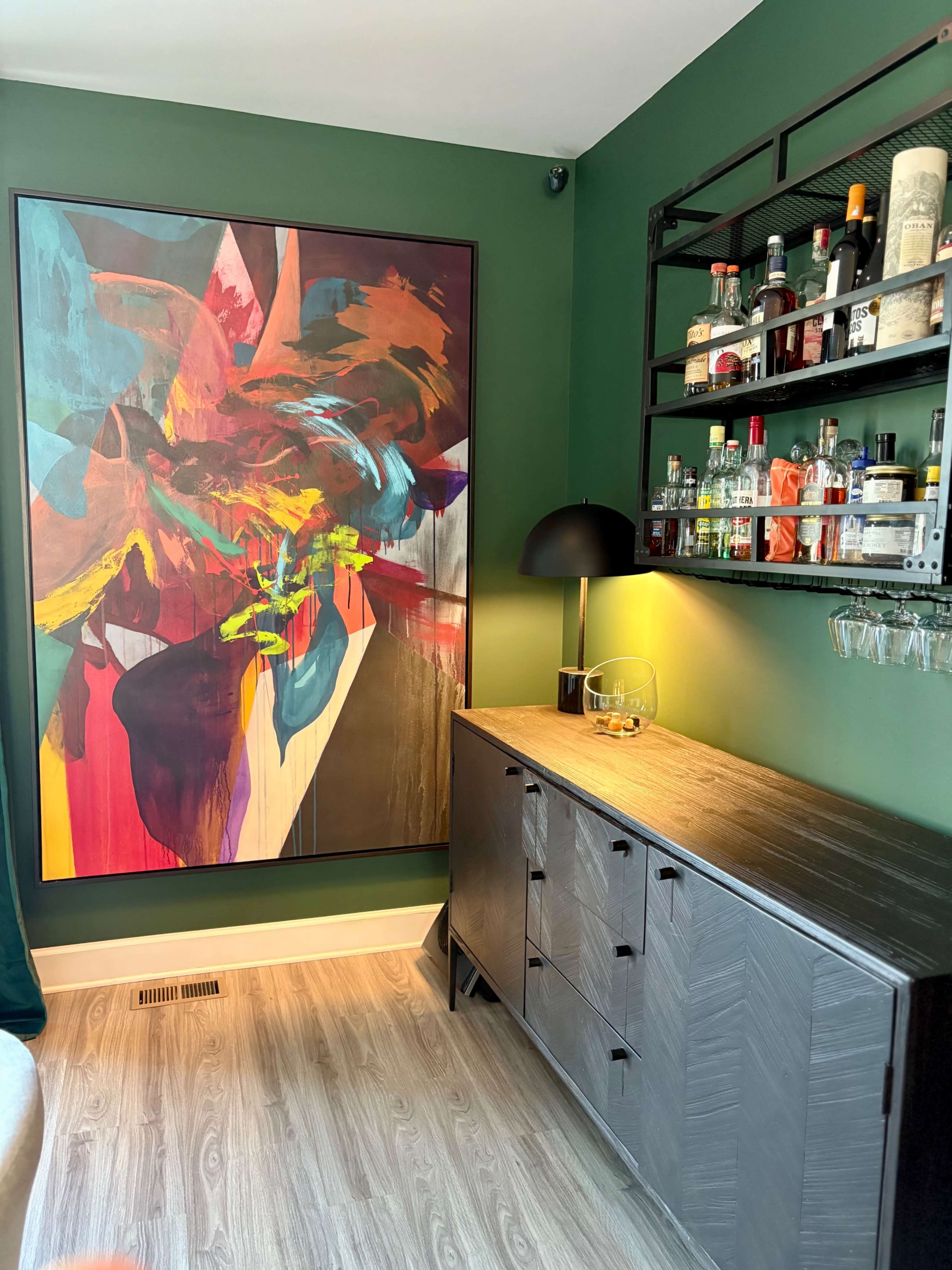 The image shows a dark green room featuring a large colorful abstract painting on one wall and a sleek bar cart with various bottles and glasses on another.