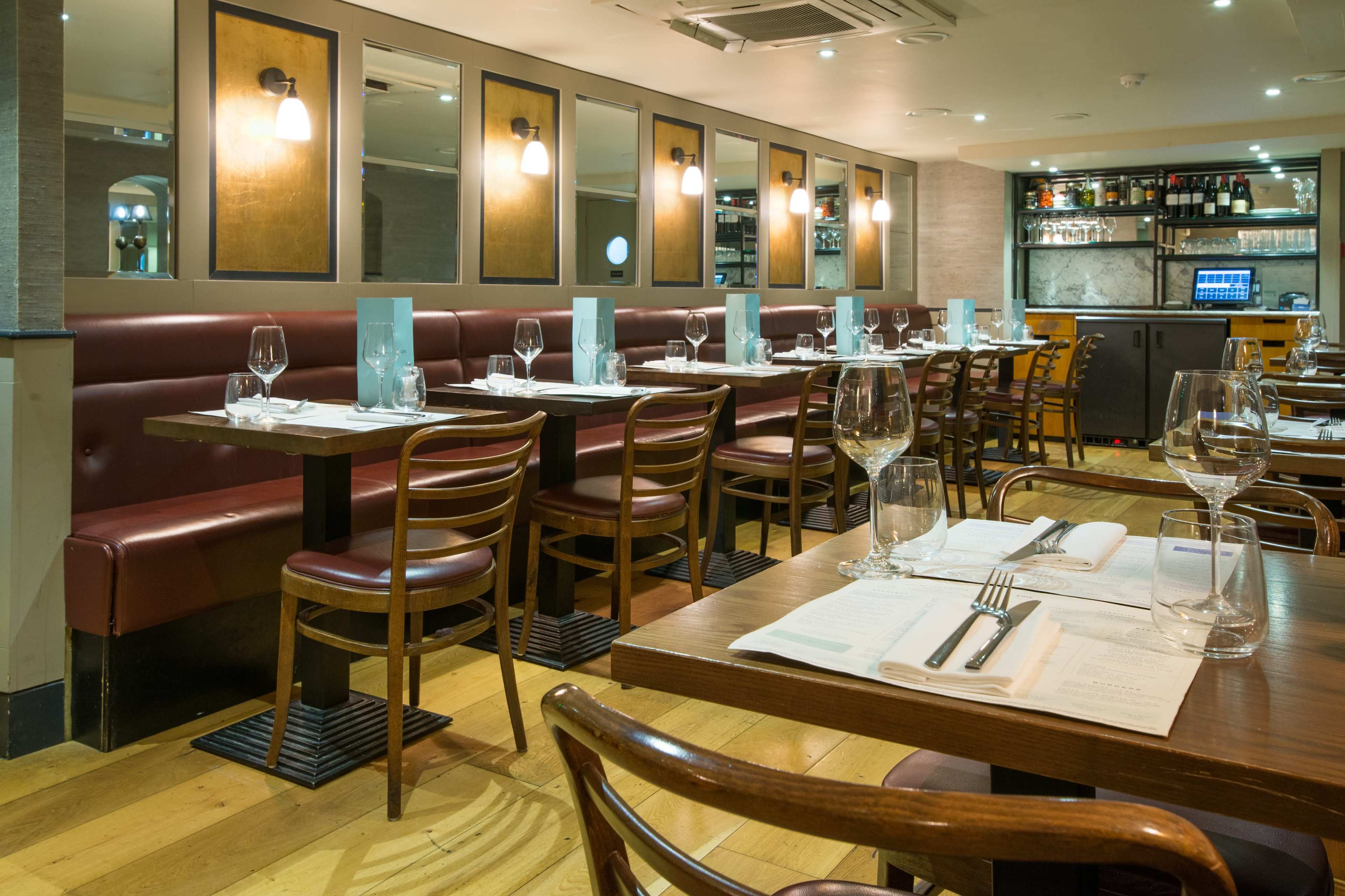 The image shows an indoor restaurant with wooden tables and chairs, red upholstered booths, and a bar in the background.