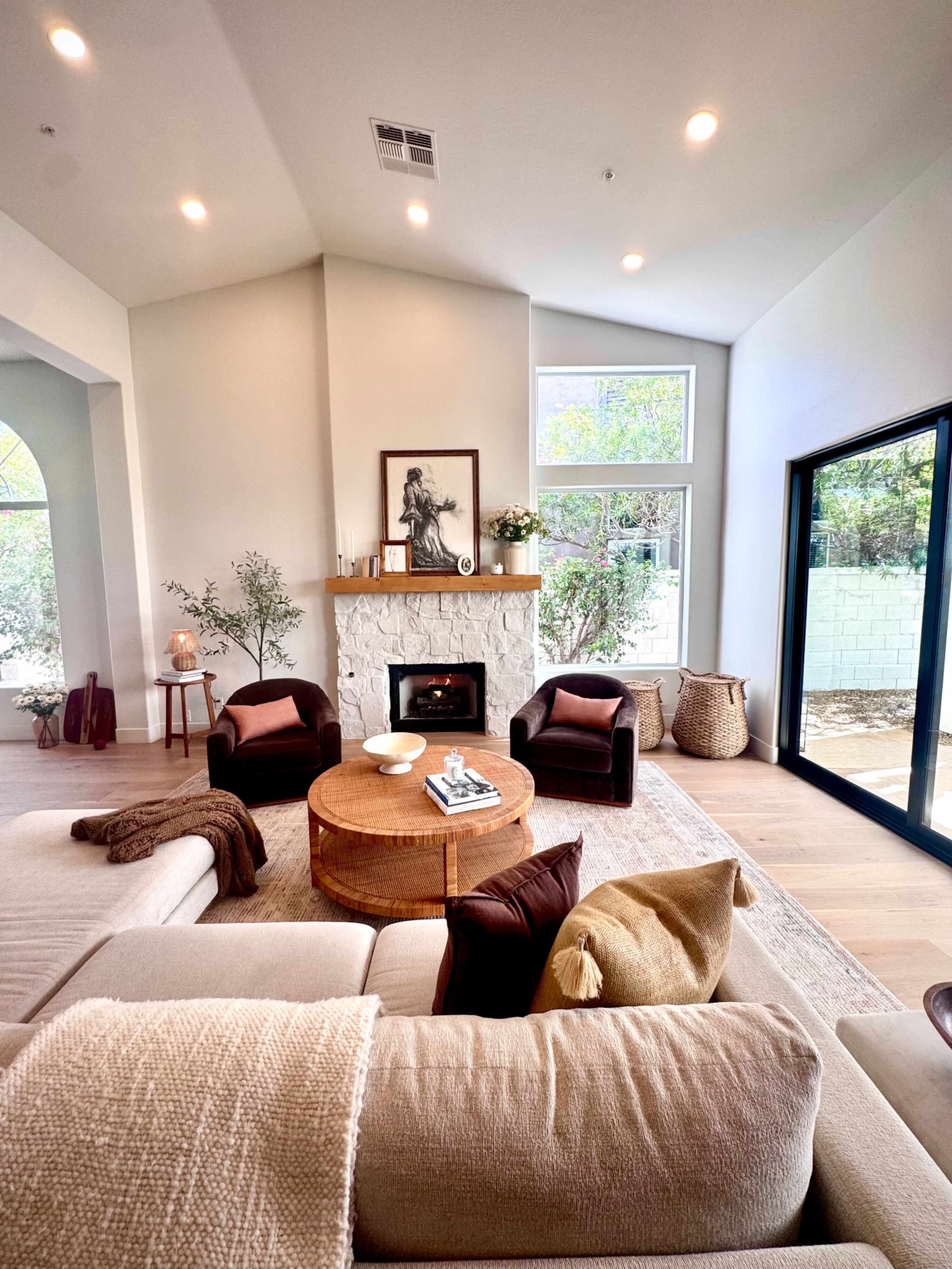 A modern living room features a stone fireplace, two dark armchairs, a round coffee table, and large windows that allow natural light to enter the space.