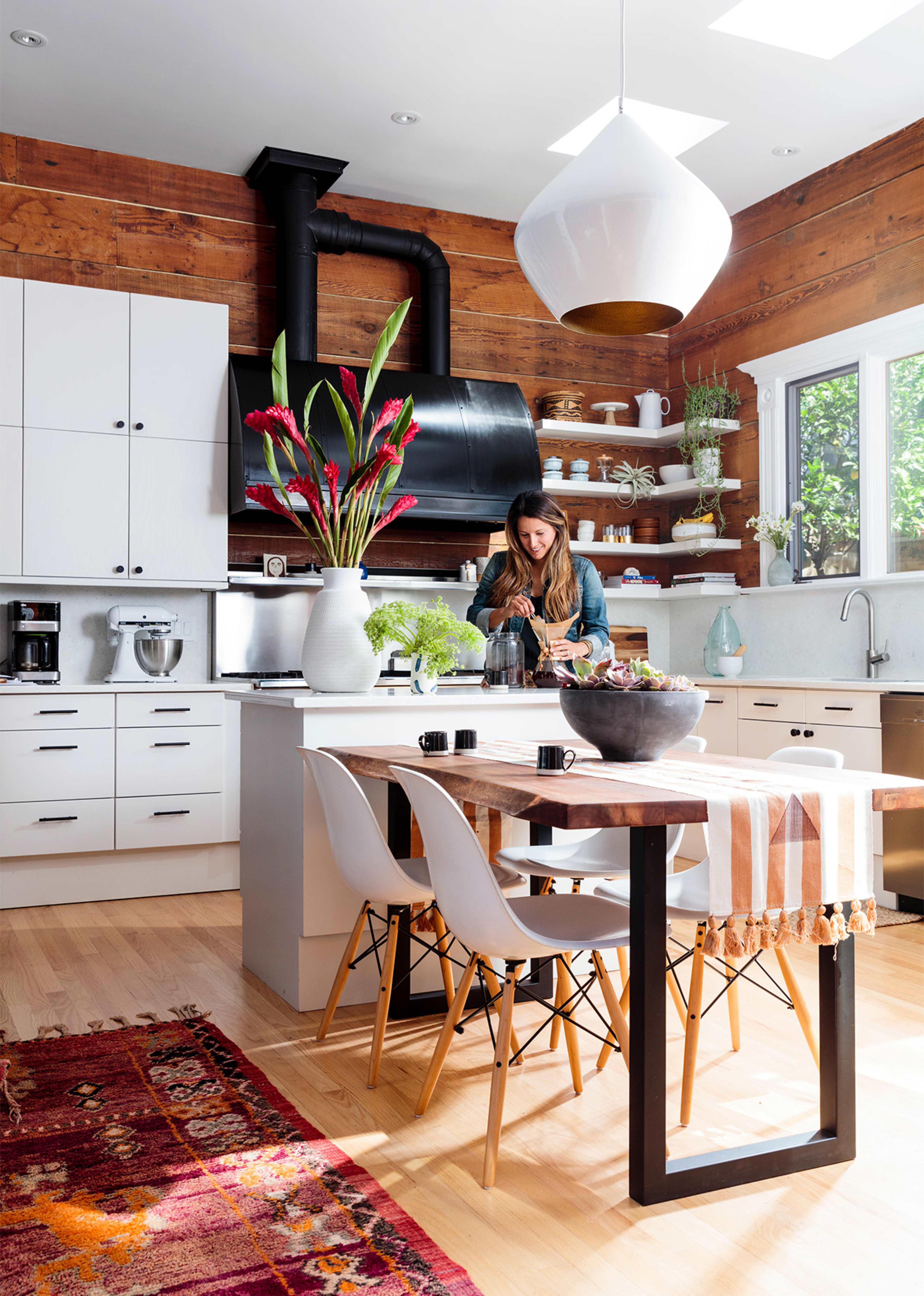 A woman prepares food in a modern kitchen with wooden walls, white cabinetry, and a large dining table.
