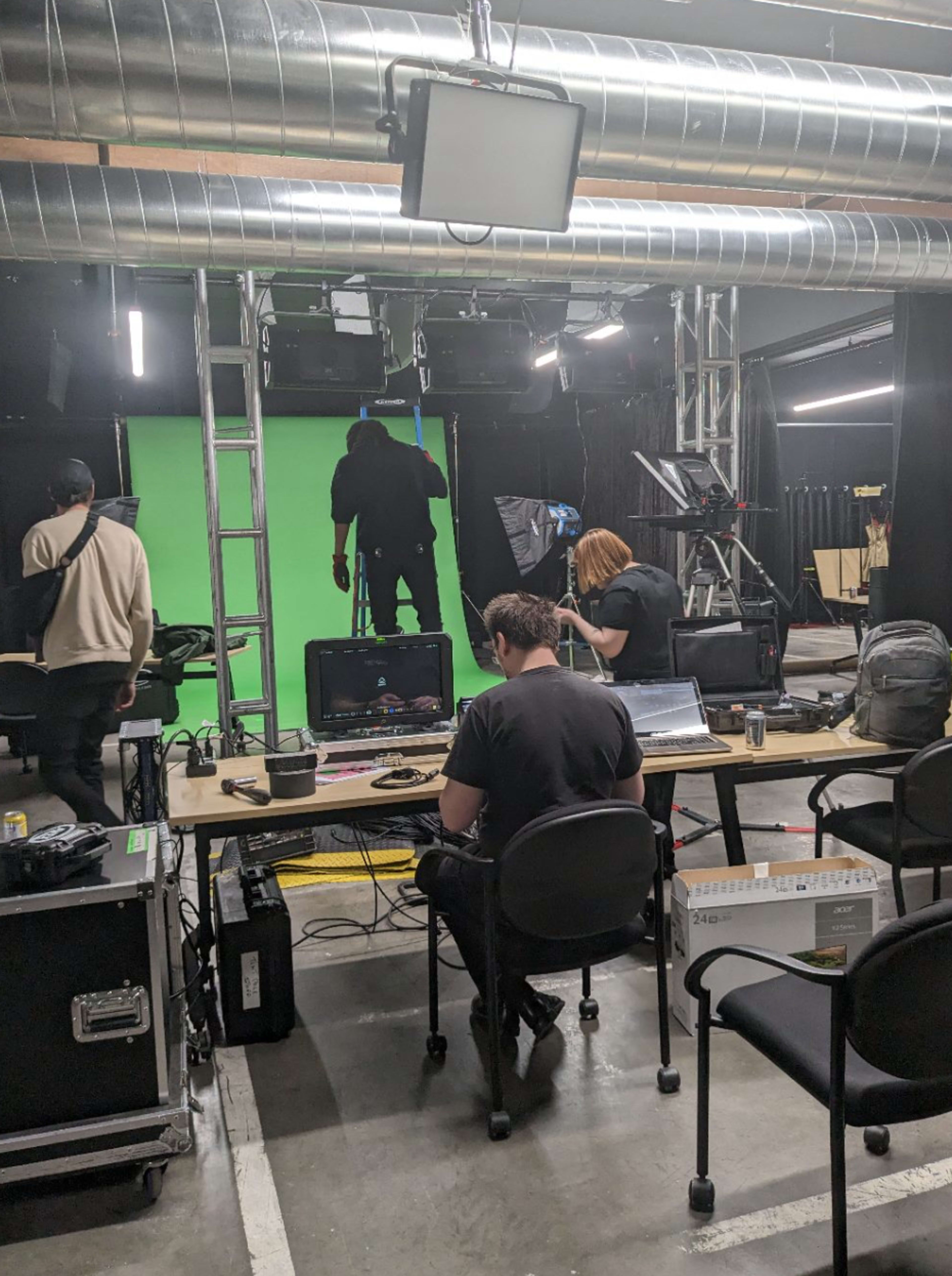 A studio set featuring a green screen, with crew members preparing equipment and working at computers.