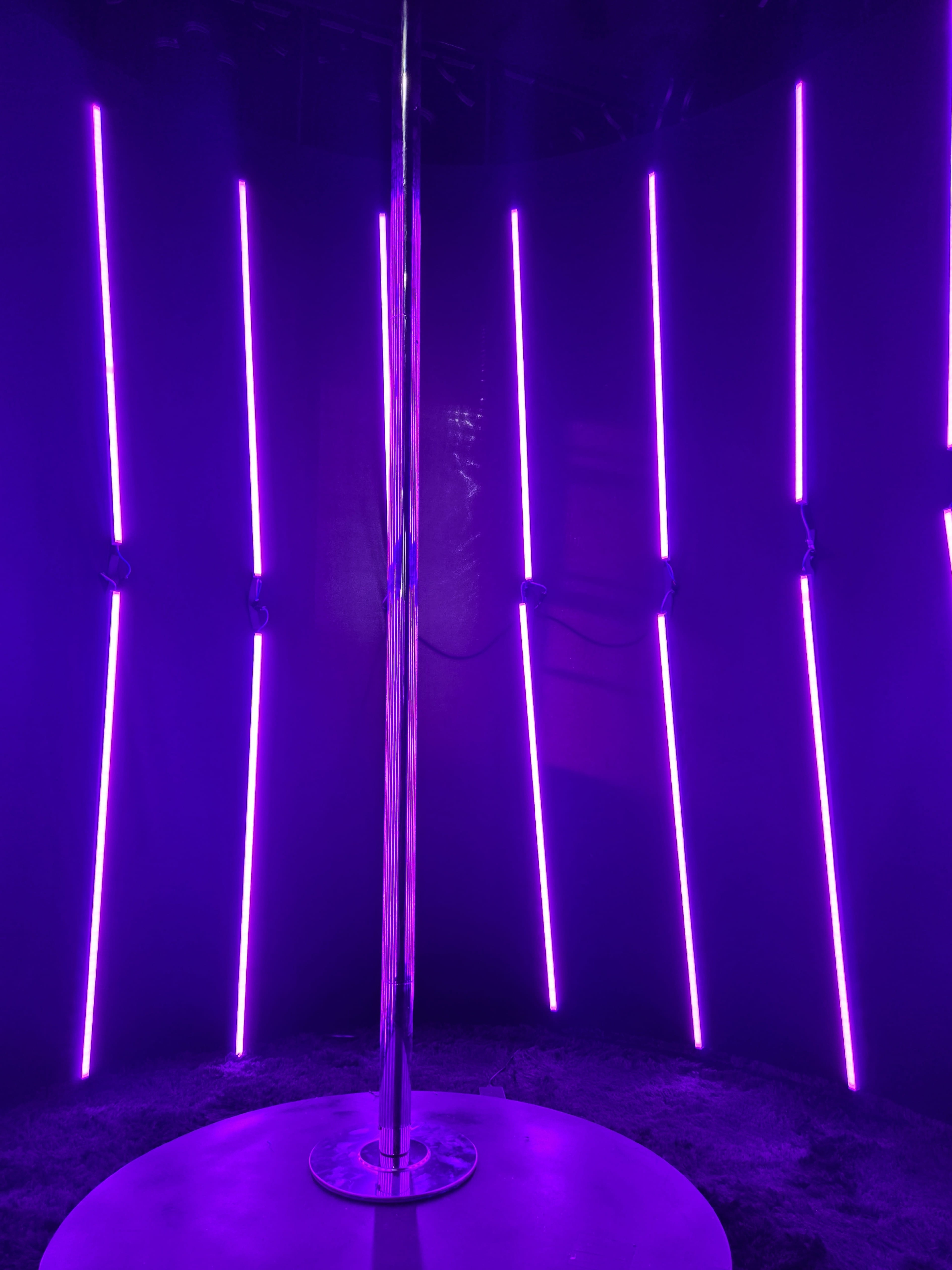 The image shows a polished metal pole surrounded by vertical neon purple lights in a dark, circular space.