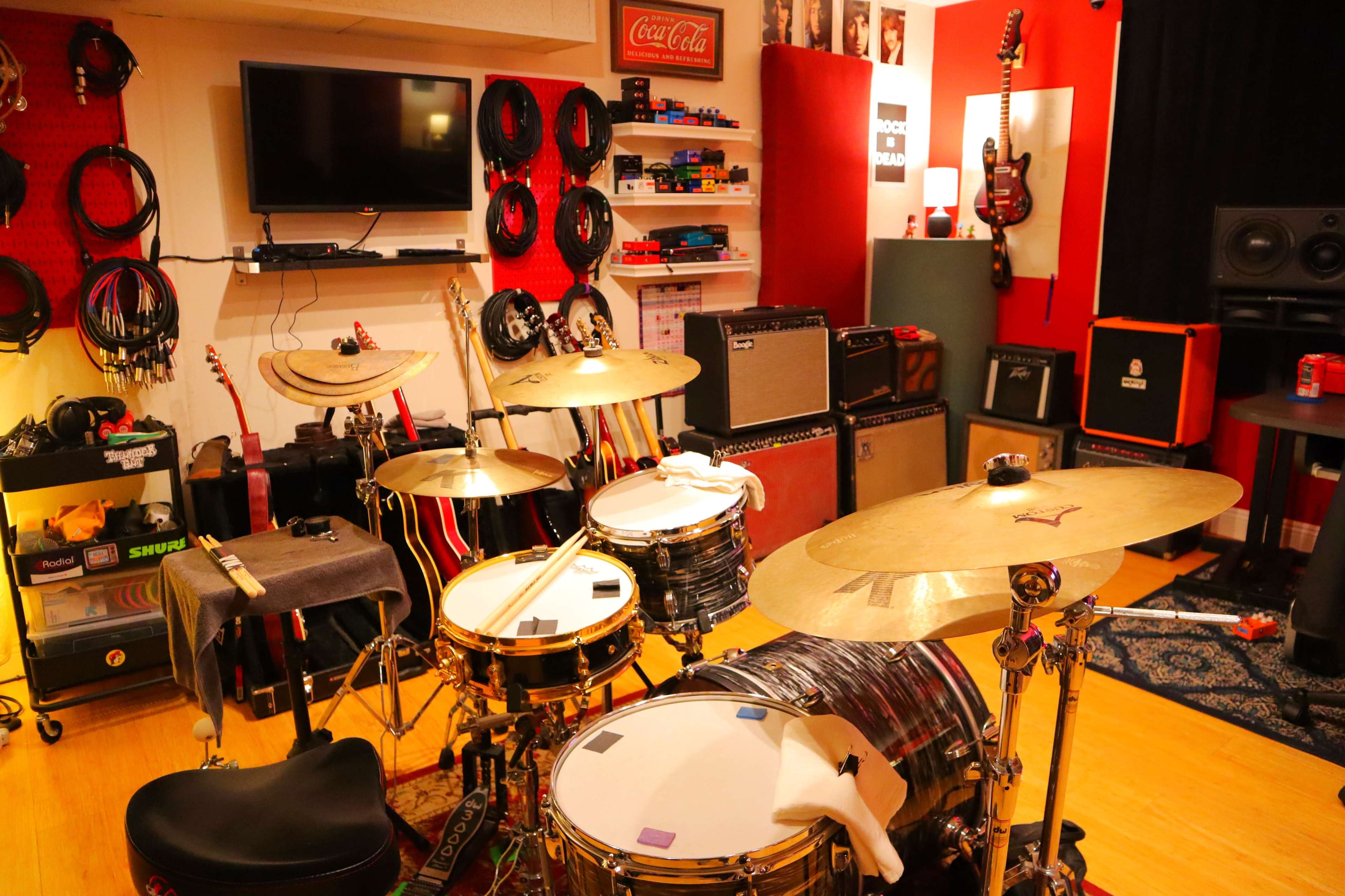 The image shows a music practice room with a drum set in the foreground, amplifiers along the walls, and various musical instruments and equipment arranged around the space.